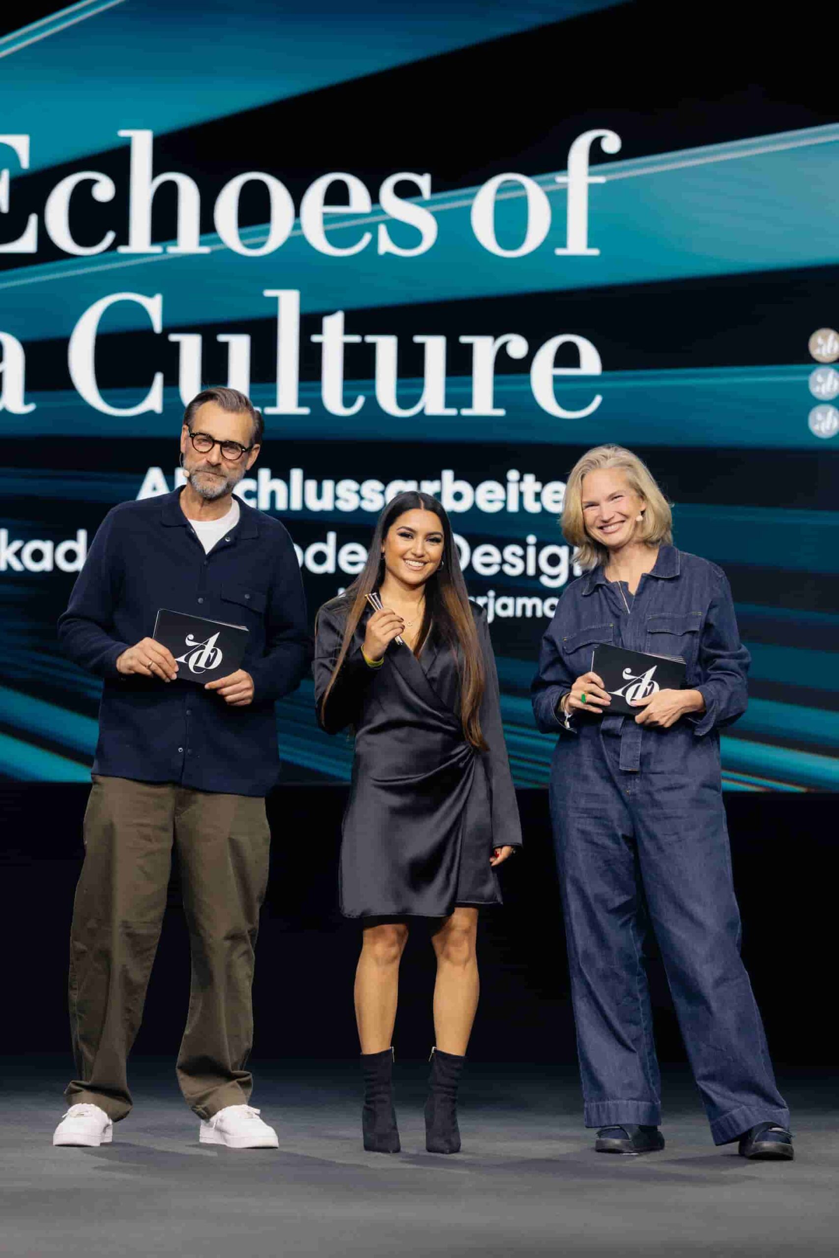 Larisa steht in der Bühnenmitte zwischen zwei Jurymitgliedern und lächelt stolz, während sie die drei Nagel-Trophäen vor einer großen Projektion präsentiert.