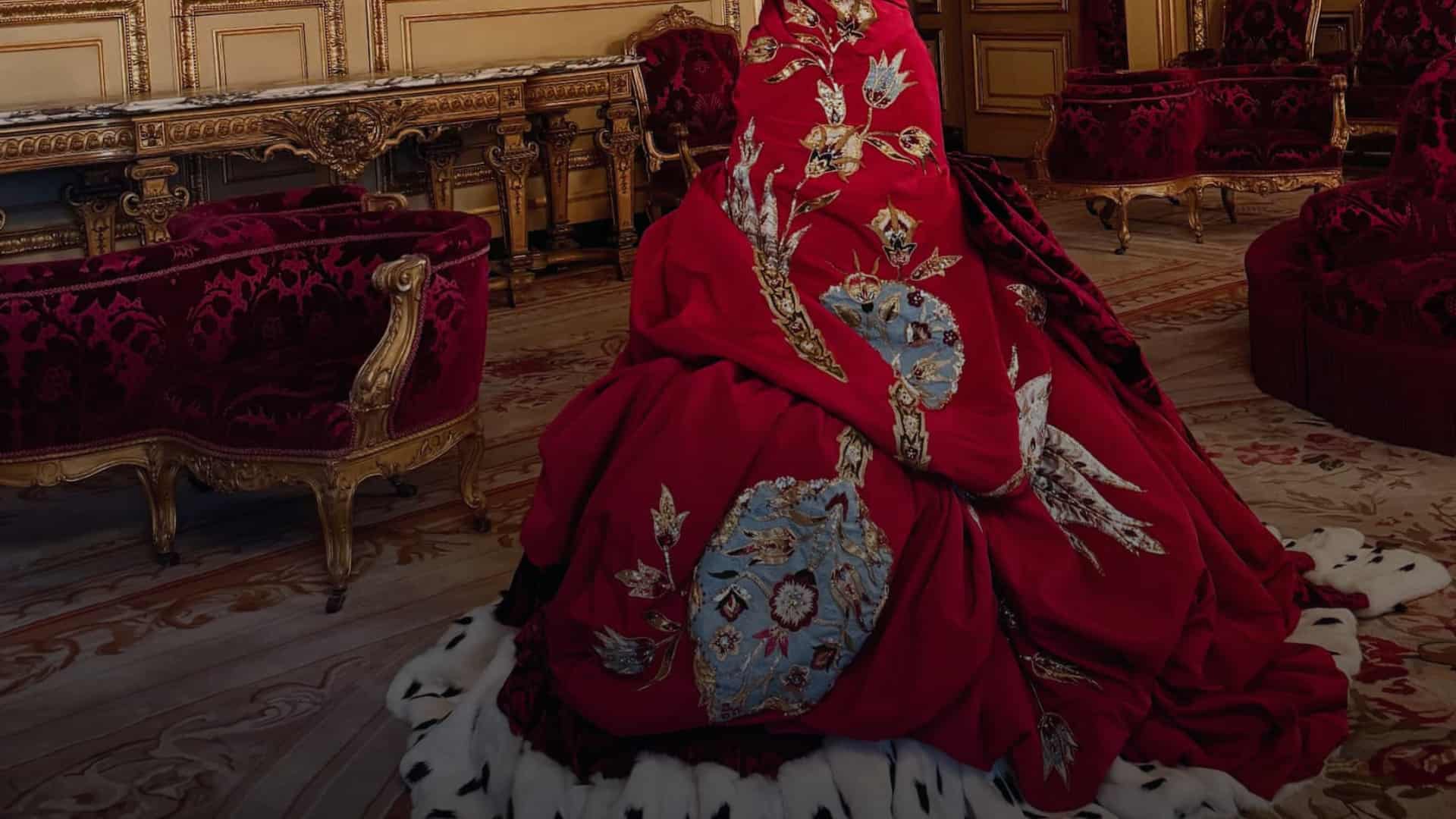 Ein elegantes Christian-Dior-Kleid in rot und weiß mit Blumen und Pfaumuster, in einem majestätischen Raum mit antiken Möbeln, großen Gemälden und prächtigen Kronleuchtern, die an einen Palast erinnern.