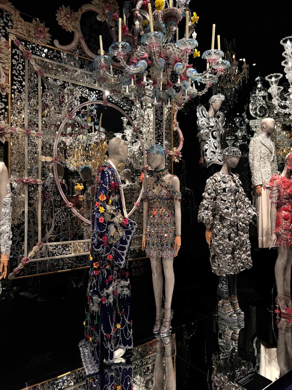 Eine fantasievolle Modenschau mit modernen Schaufensterpuppen, die besondere Kleider von Dolce & Gabbana tragen, in einem Raum mit spiegelndem Boden, großem Spiegel und glitzernden Kronleuchtern. Dominierend silberne Optik mit dezenten Farbtupfern.