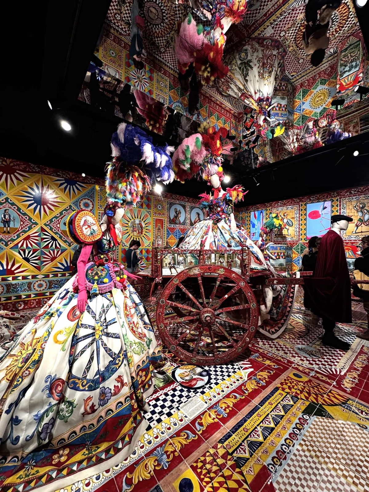 Eine farbenfrohe Kunstinstallation mit bunten Dolce & Gabbana-Kleidern, einer spiegelnden Decke und einem Wagen im Hintergrund, auf dem ein Kleid präsentiert wird.