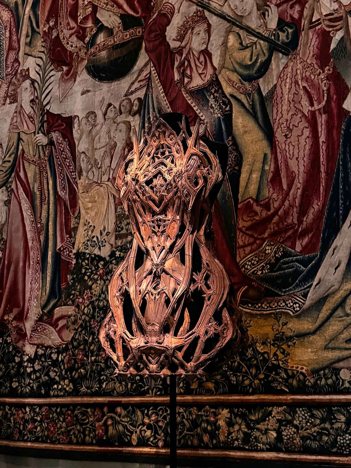 Futuristische 3D-Print-Skulptur eines Kleides mit verschiedenen Verzierungen, die an einen Science-Fiction-Film erinnern, vor einem historischen Wandteppich als Hintergrund.