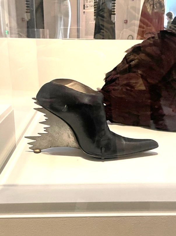 Ein Designer-Schuh im Art-déco-Stil mit Metallelementen und schwarzem Leder, ausgestattet mit einem auffälligen, kreativen Absatz. Der Schuh wird in einer Vitrine mit Lichtspot präsentiert und zieht alle Blicke auf sich.