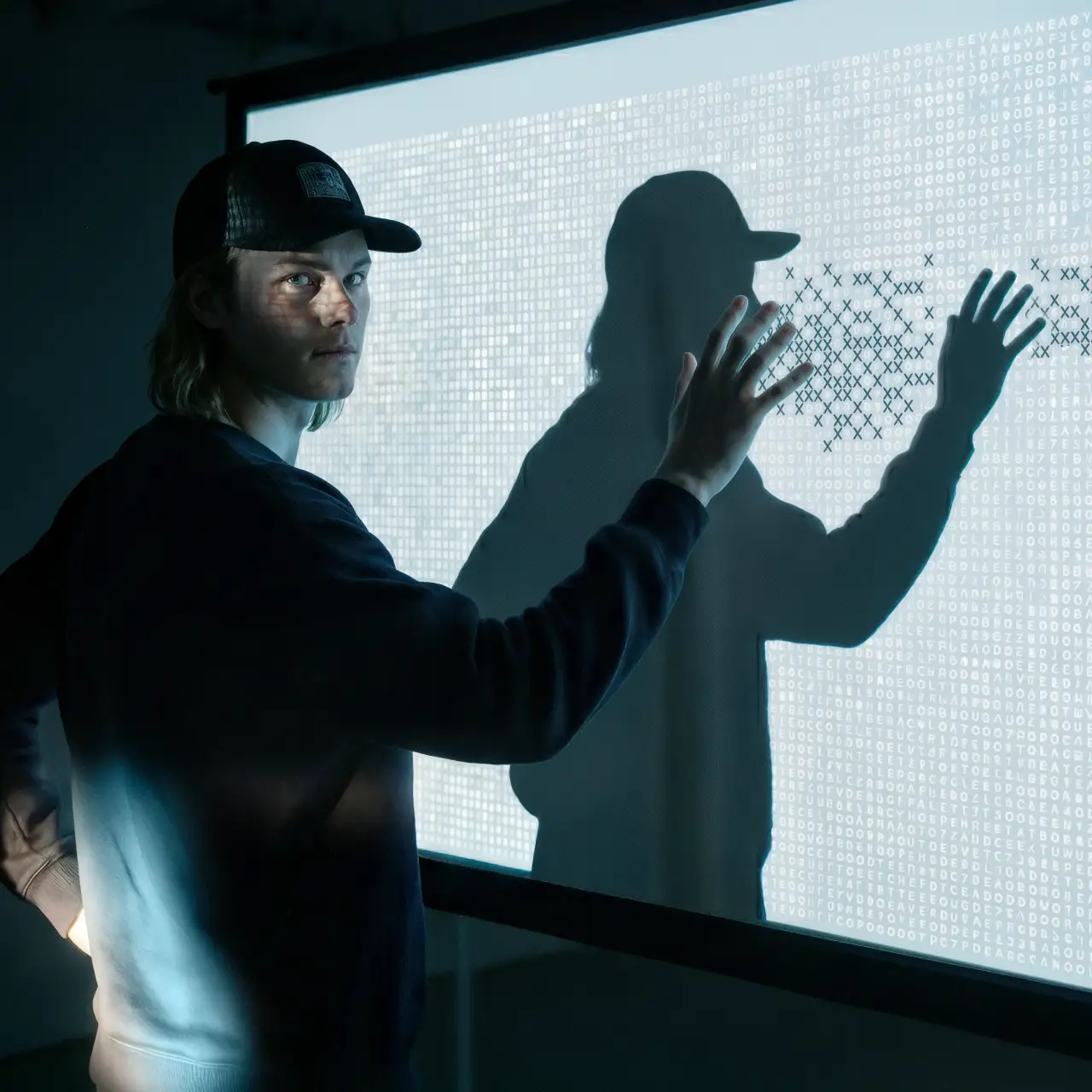 Ein junger Mann mit Baseballkappe blickt nach vorn und berührt mit der Hand eine digitale Oberfläche, die auf eine Leinwand projiziert wird.