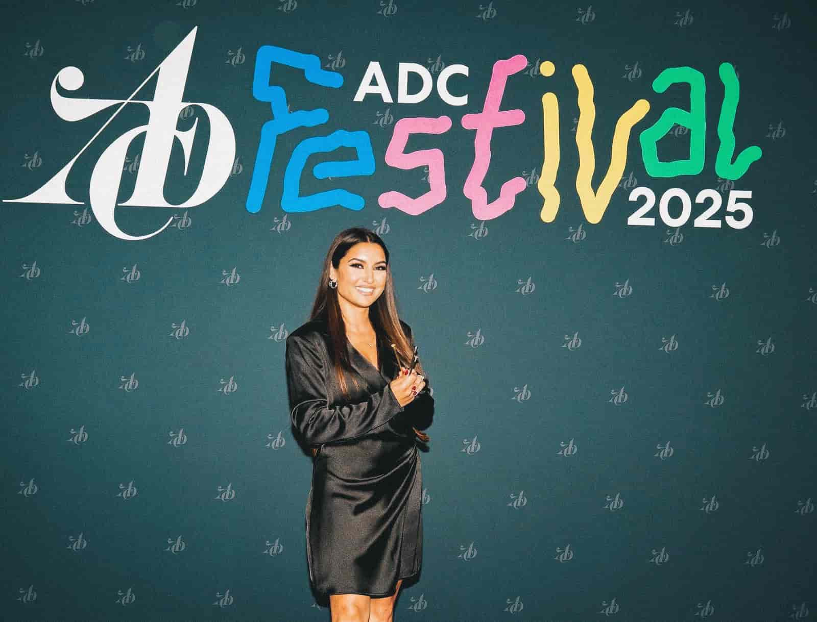 Larisa Barjamovis lächelt, während sie vor einem Plakat mit dem ADC Festival 2025 Logo posiert und die drei Nägel-Trophäen in den Händen hält.
