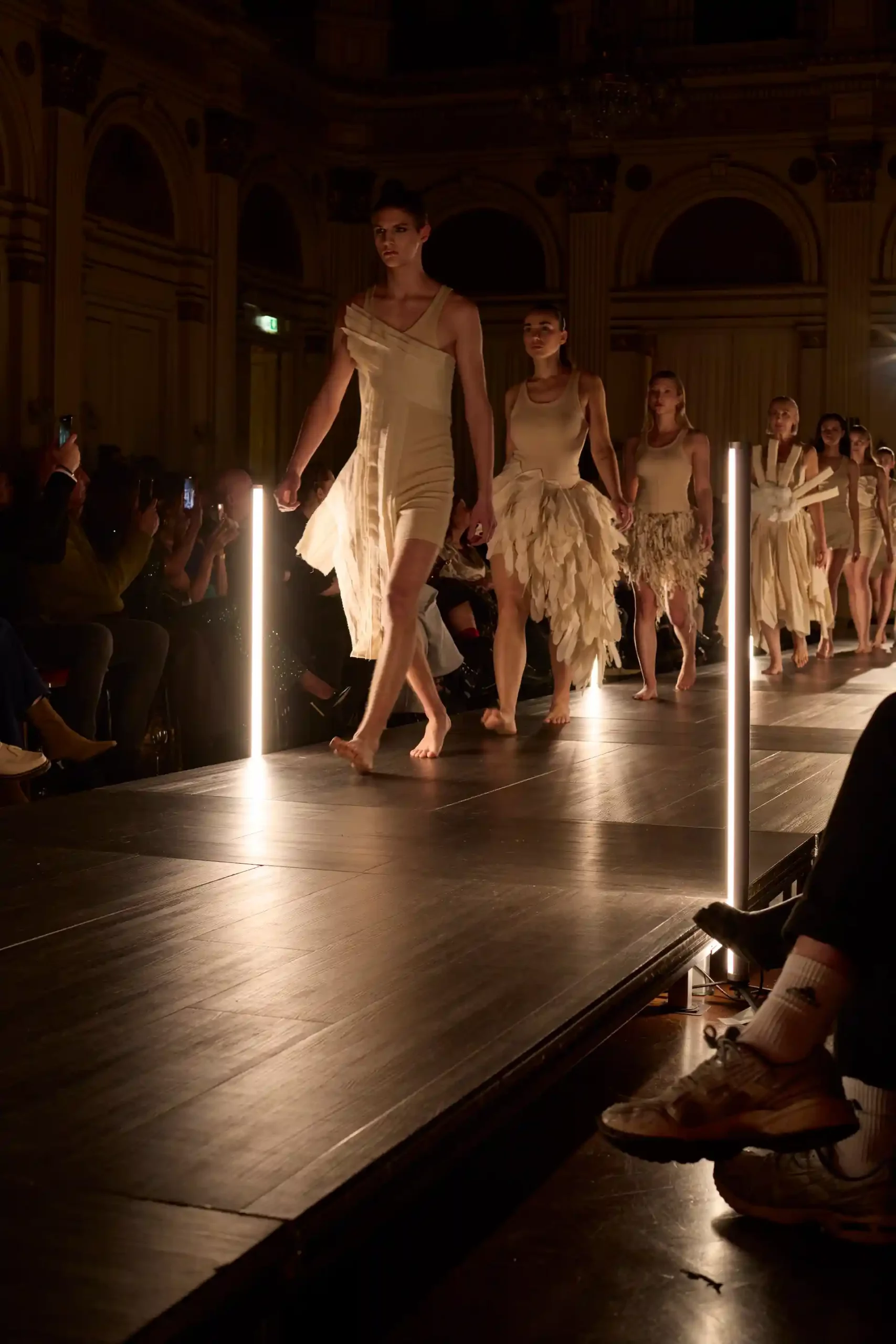 Mehrere Models schreiten barfuß in sandfarbenen, futuristischen Kleidern über den Laufsteg, während der Saal dunkel bleibt und lediglich der Laufsteg von seitlichen Scheinwerfern erhellt wird.
