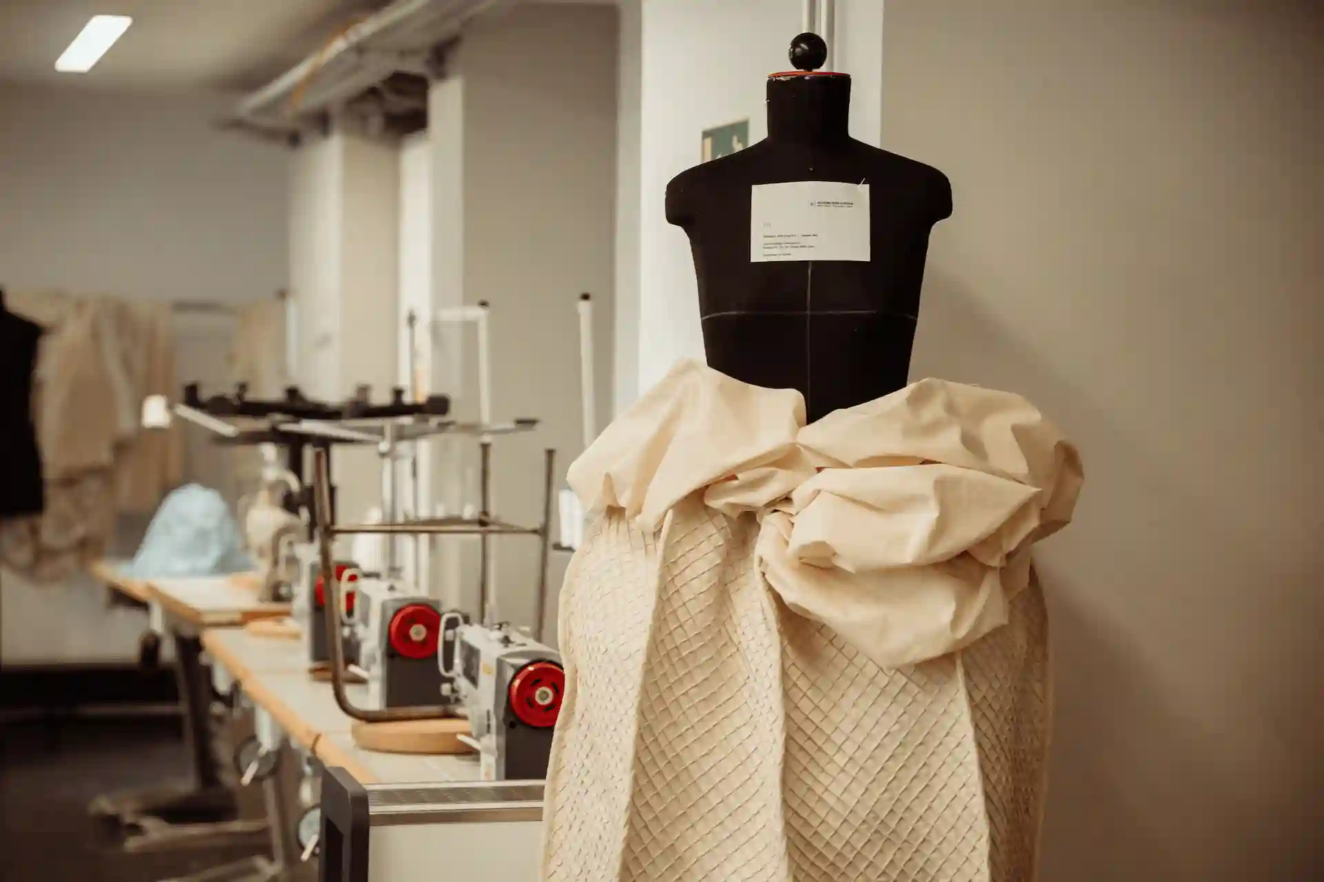 Schwarze Schneiderbüste mit hellbeigem, drapiertem Stoff; im Hintergrund Nähmaschinen und Arbeitsplätze in einem Atelier.
