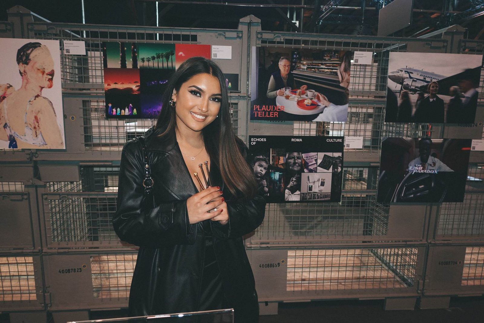 Larisa, in schwarzer Kleidung, hält lächelnd und stolz ihre drei Nagel-Trophäen. Im Hintergrund sind ihre Arbeiten an einer Wand ausgestellt.