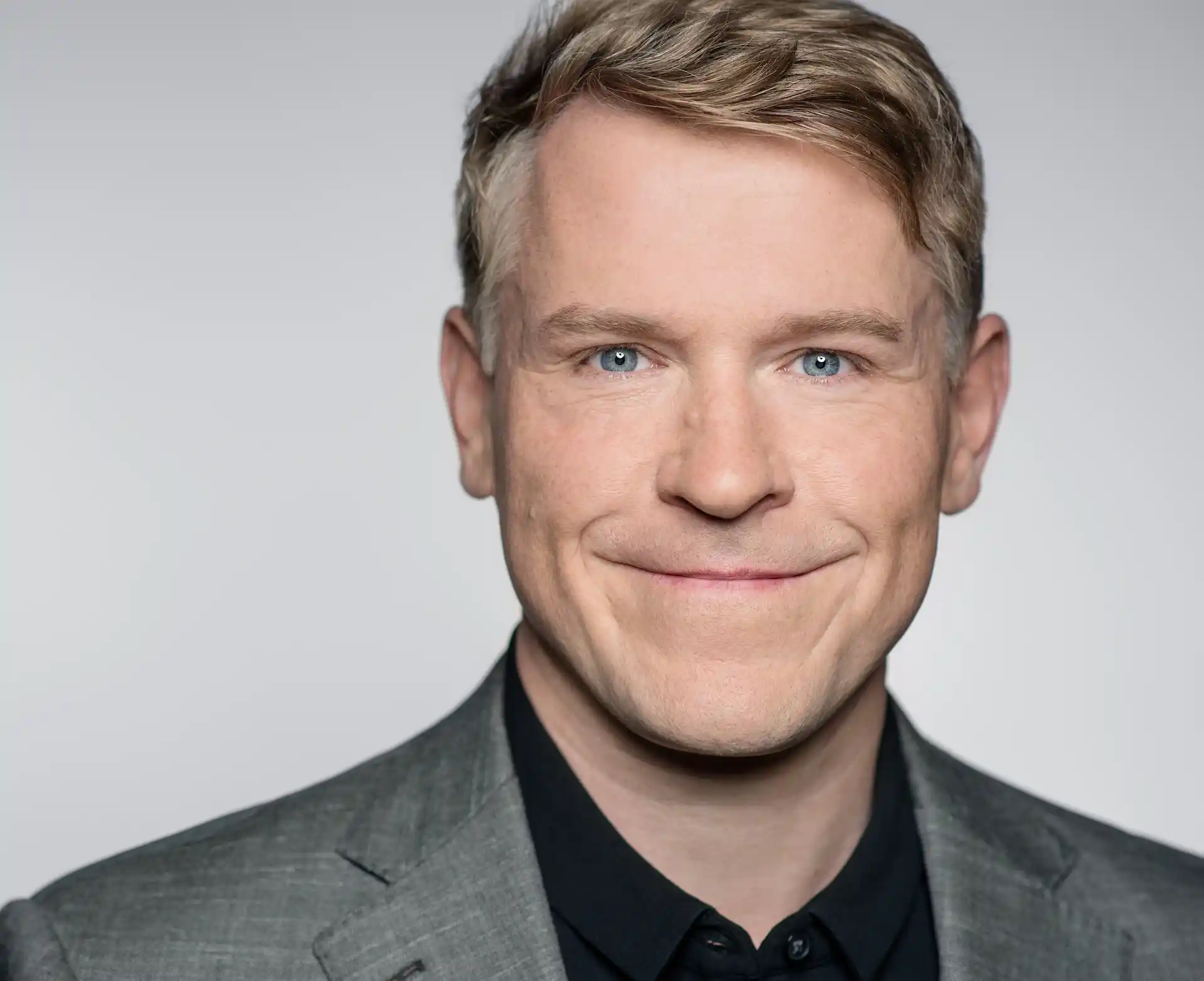 Portrait von Prof. Rollwagen, der in die Kamera lächelt, gekleidet in ein graues Sakko und ein schwarzes Hemd, mit blondem Haar und blauen Augen.