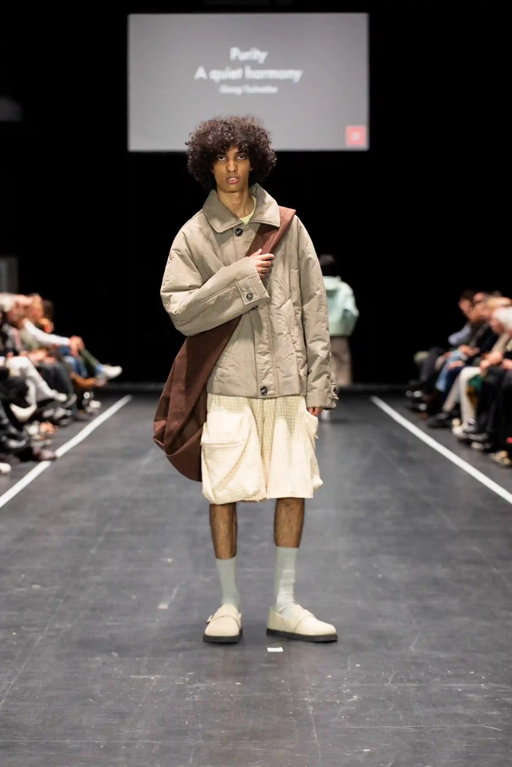 Ein Color-of-People Mann mit lockigem Haar steht auf dem Laufsteg. Er trägt eine modische Jacke, kurze weiße Hosen und einen langen braunen Rucksack.