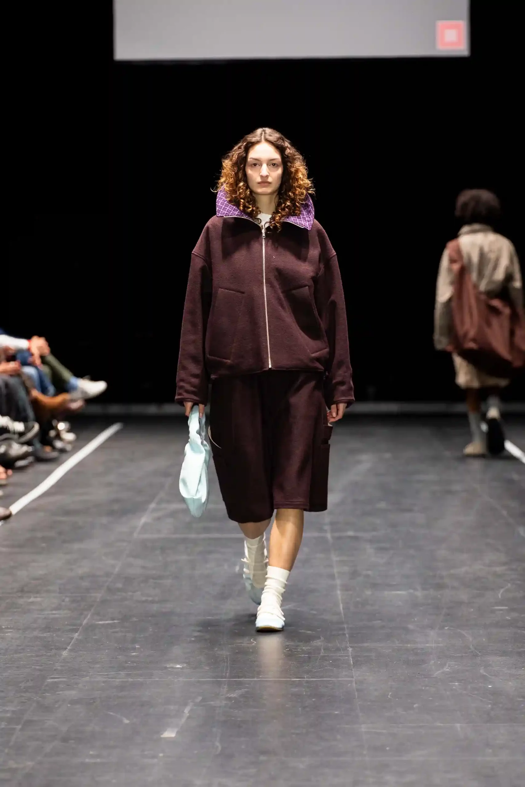 Model auf dem Laufsteg mit lila Kragen, kurzer Hose, weißen Turnschuh und braunem Outfit.