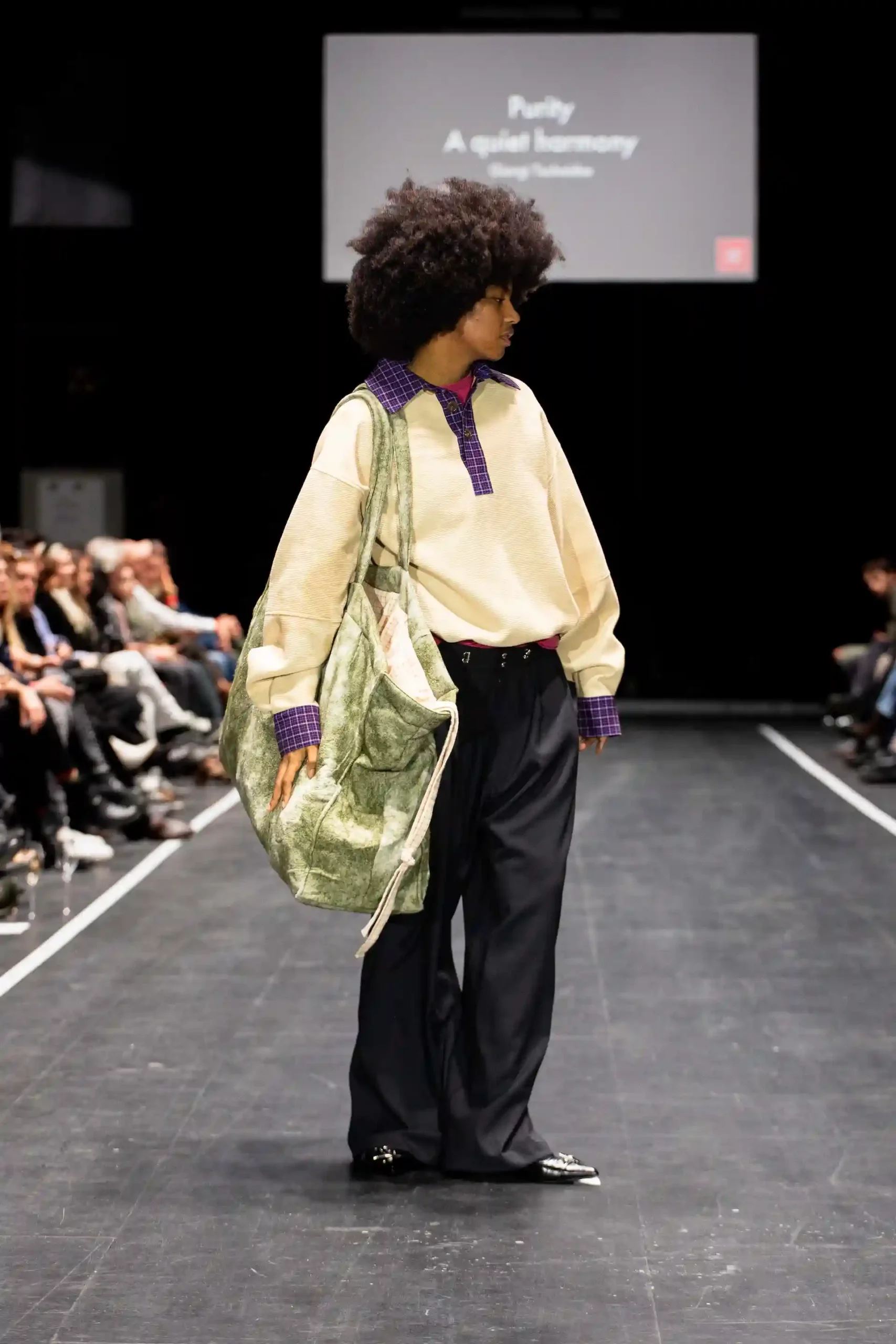 Laufstegmode mit einem People of Color Model, das ein auffälliges Outfit und eine große Tasche trägt. Sie hat eine Afrofrisur wie aus den 70ern, trägt einen cremefarbenen Pulli, eine weit geschnittene schwarze Hose und glänzende schwarze Schuhe.