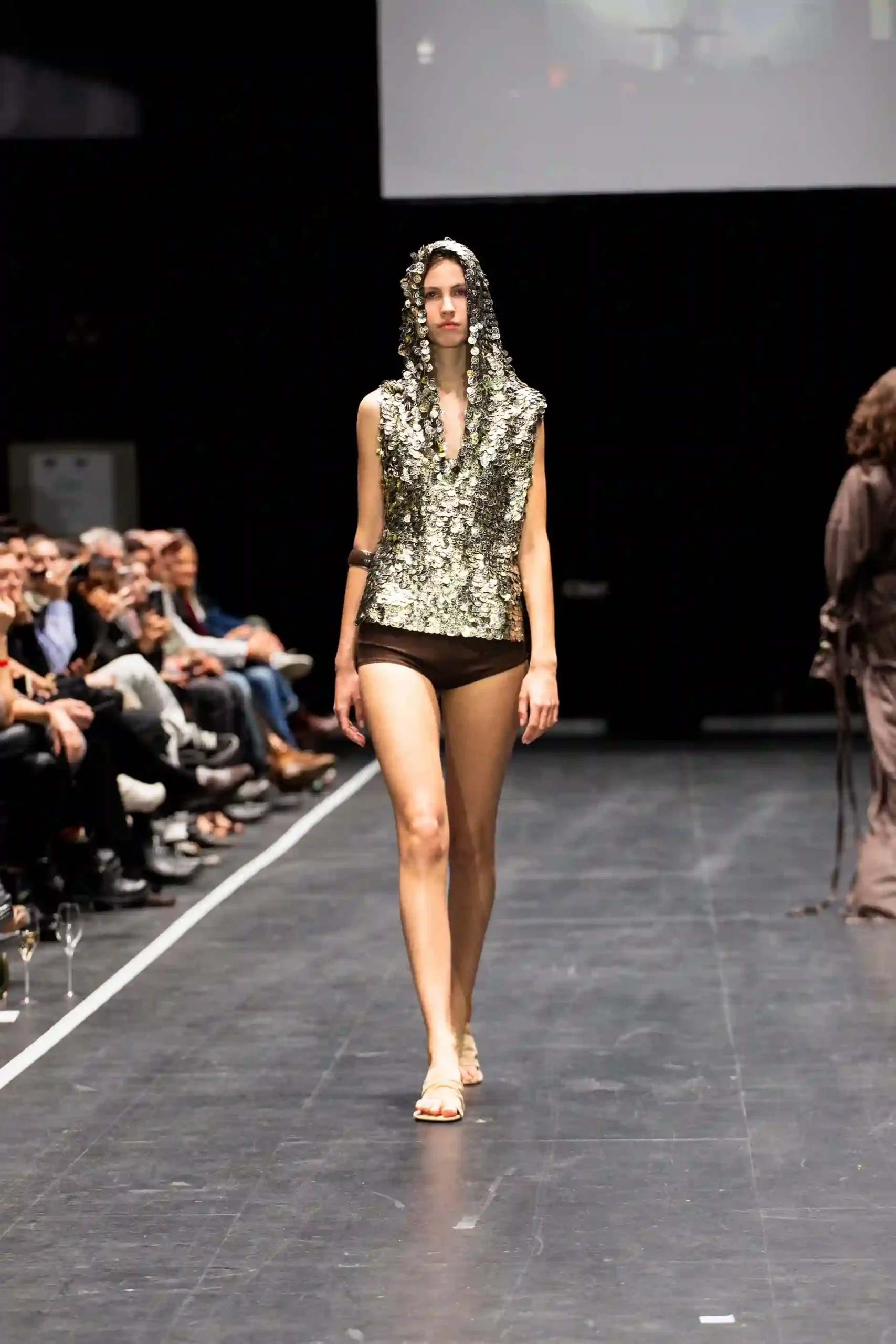 Model auf der Modenschau mit goldenem Oberteil und Kapuze und braune Shorts