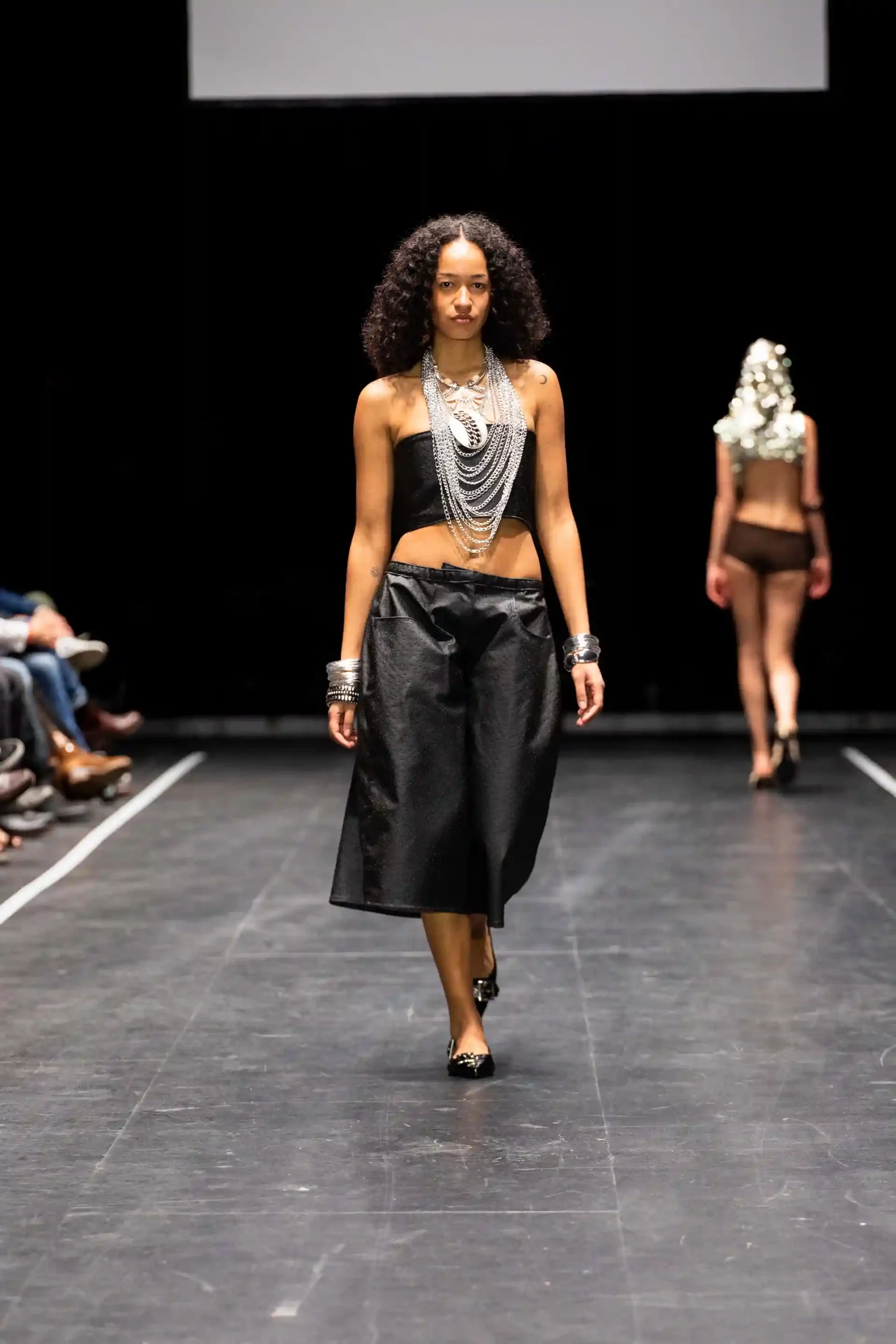 Eine Modenschau mit einem People-of-Color-Model in schwarzem Rock und auffälligem Schmuck: einer langen Kette und silbernen Armreifen.