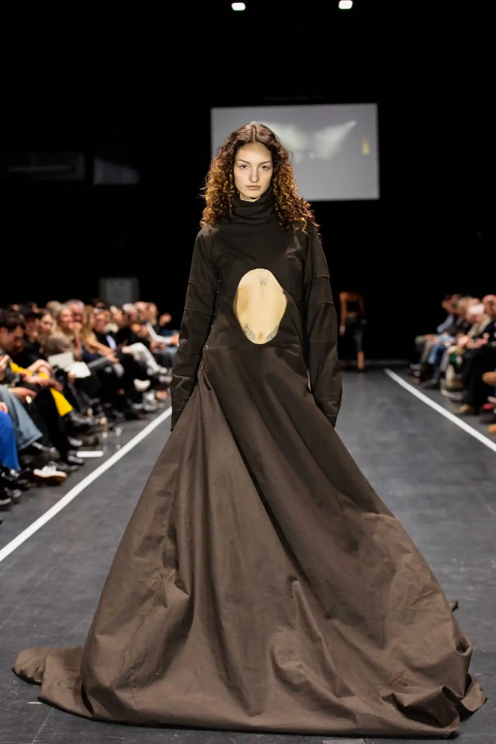 Model mit lockigem Haar trägt ein stilvolles braunes Kleid mit einzigartigem Design auf dem Laufsteg.