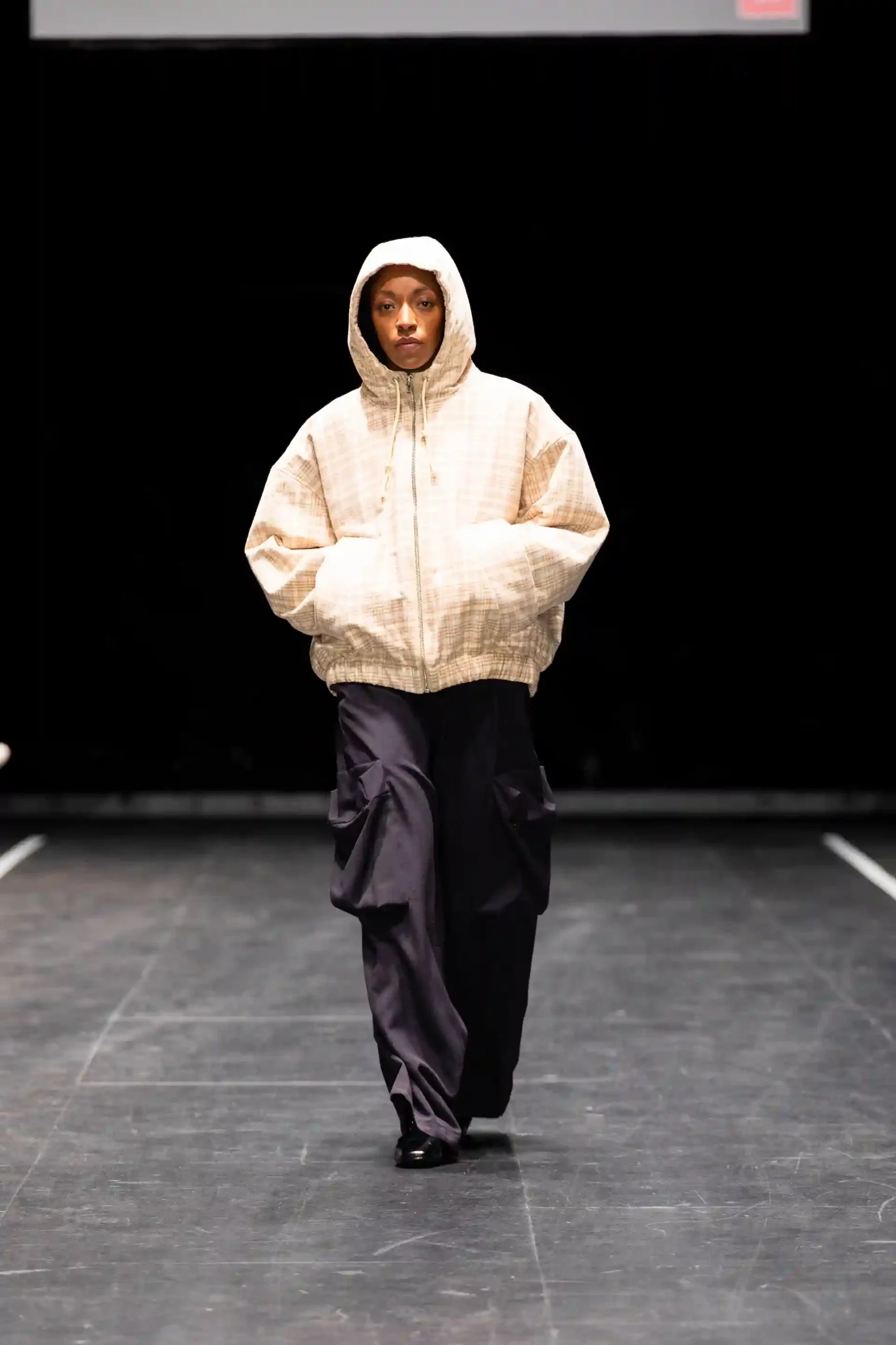 Model in einer beigen Jacke, mit Kaputze und schwarzen Hosen auf dem dunklem Laufsteg.