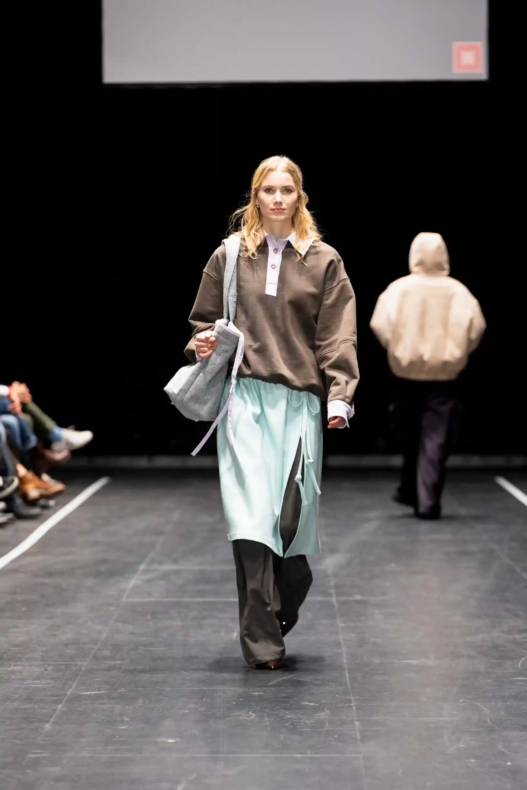 Model auf dem Laufsteg in einem braunen Sweatshirt, mintgrünem Rock und hellblauer Tasche.