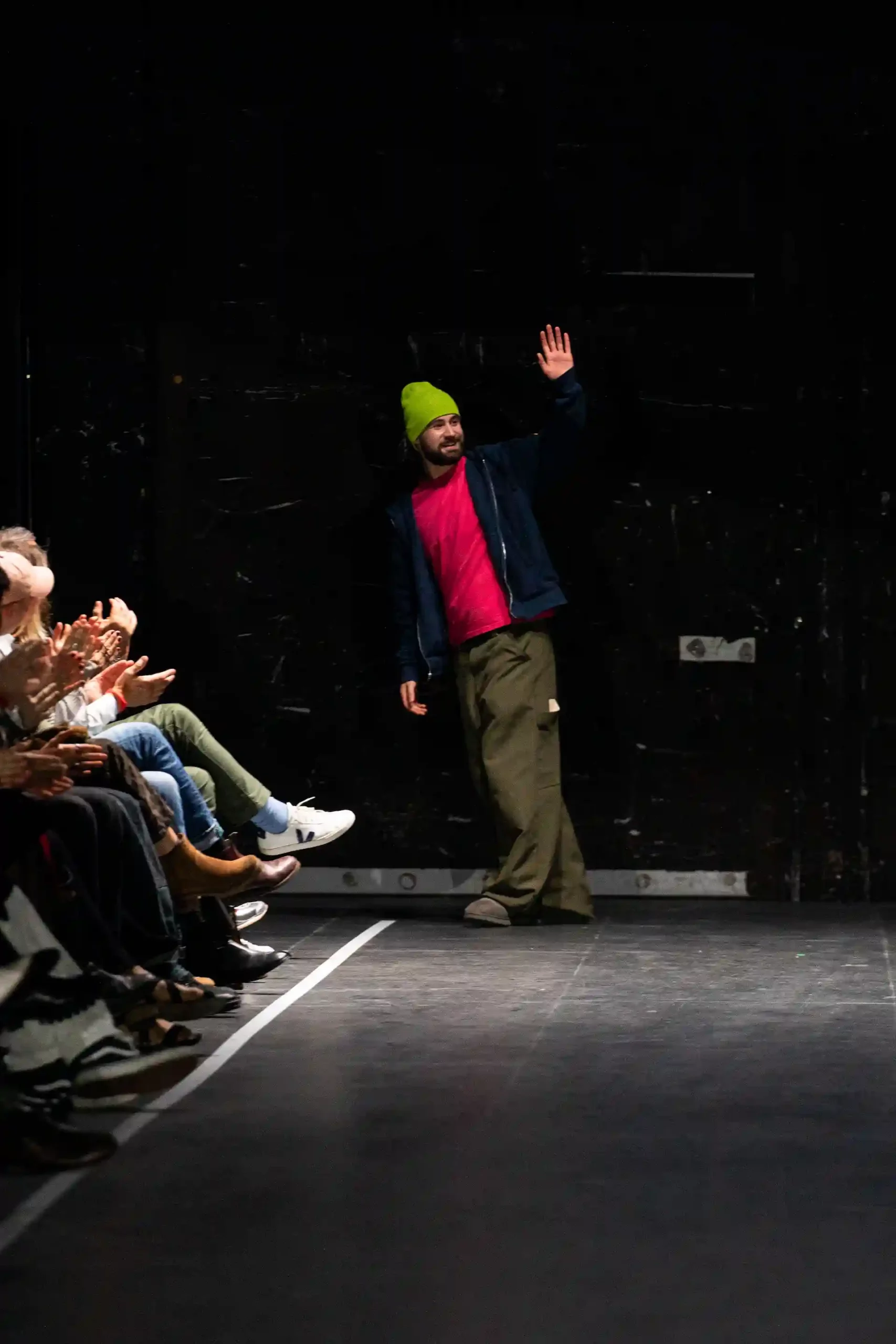 Ein Designer winkt dem Publikum auf der Bühne während die Zuschauer applaudieren.