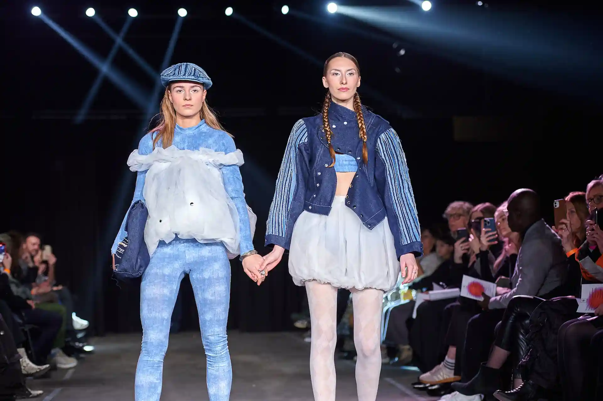 Zwei Models präsentieren modische Denim-Outfits auf dem Laufsteg während das Publikum mit ihren Handys fotografiert.