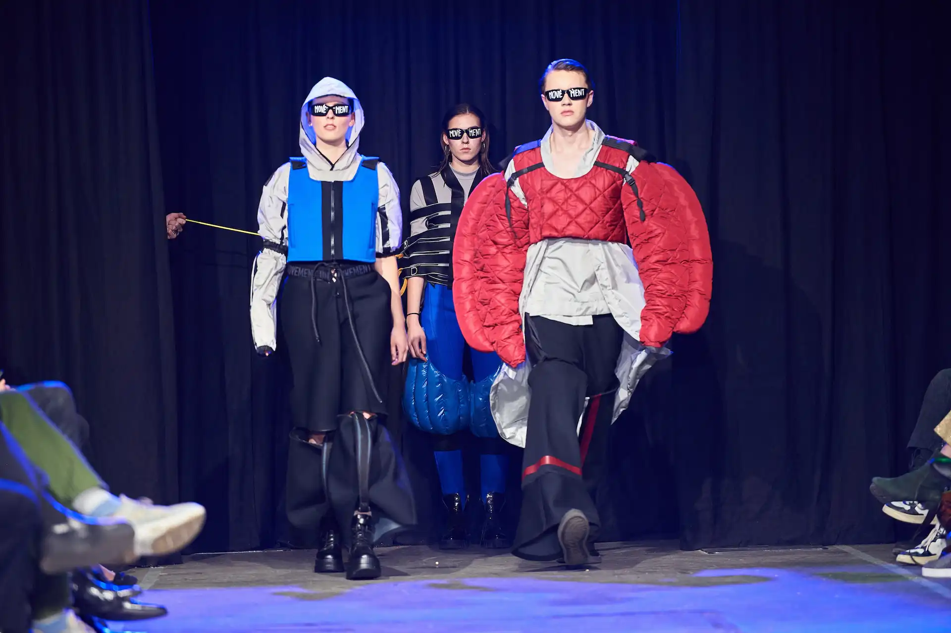 Drei Models gehen über den Laufsteg und präsentieren extrem schräge Outfits und Brillen mit Aufschrift.