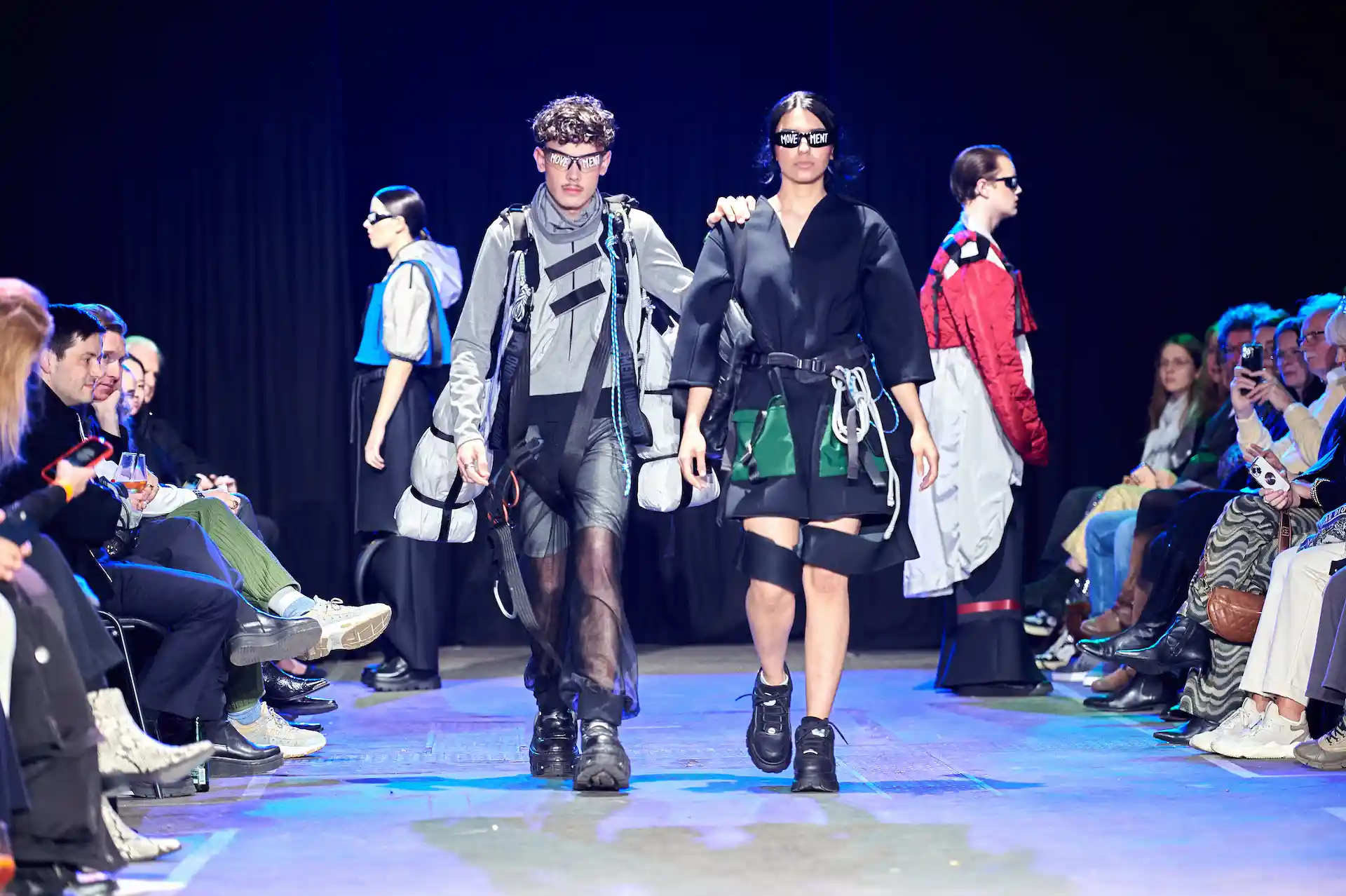 Vier Models laufen über den Laufsteg und präsentieren sehr auffällige Kleider, besondere Outfits und Brillen mit Aufschrift.