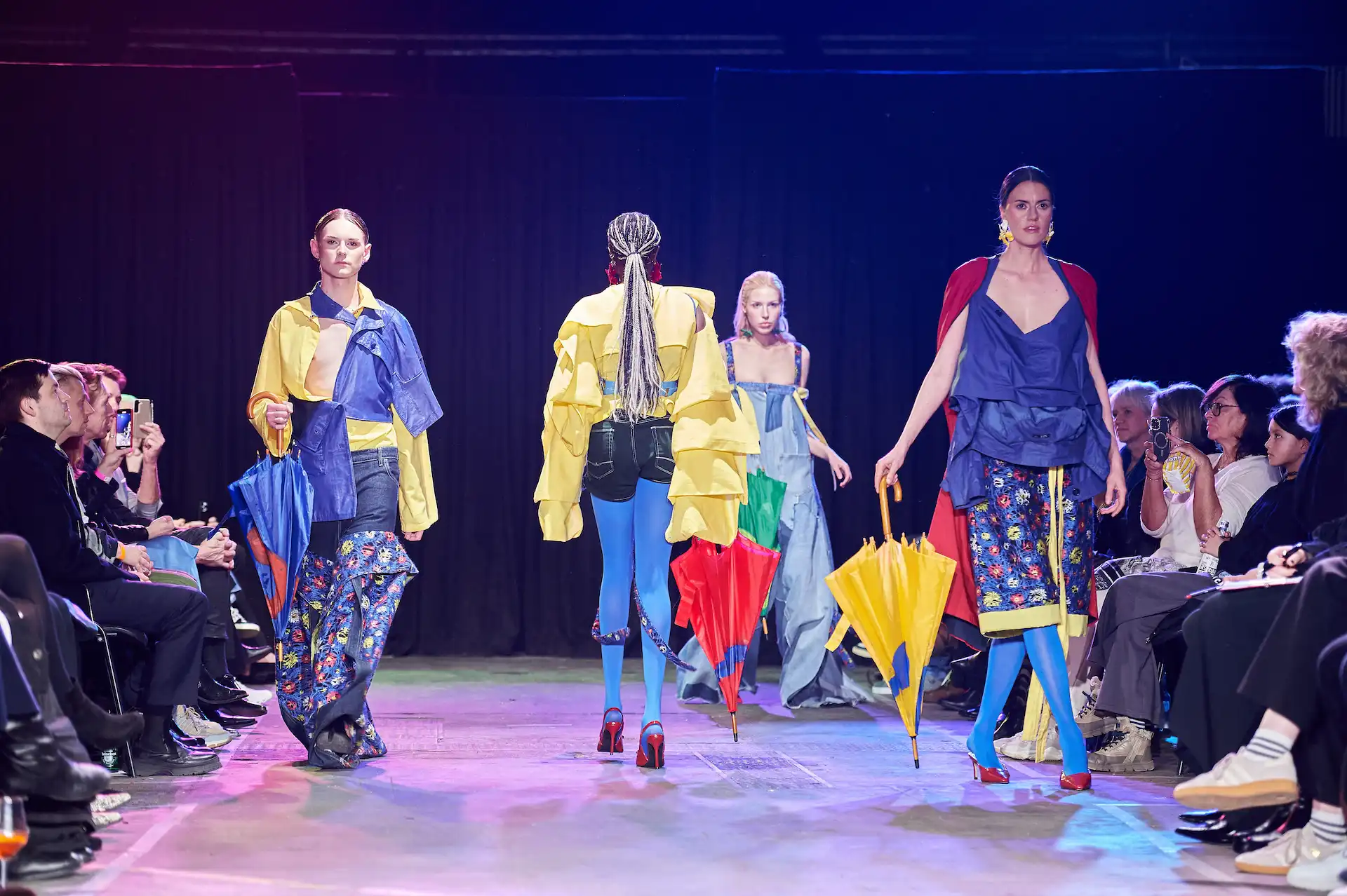 Vier Models auf dem Catwalk präsentieren sehr bunte Kleider mit Accessoires; es wirkt wie aus einem Marvel-Comic. Die Zuschauer sind begeistert und fotografieren mit ihren Handys.