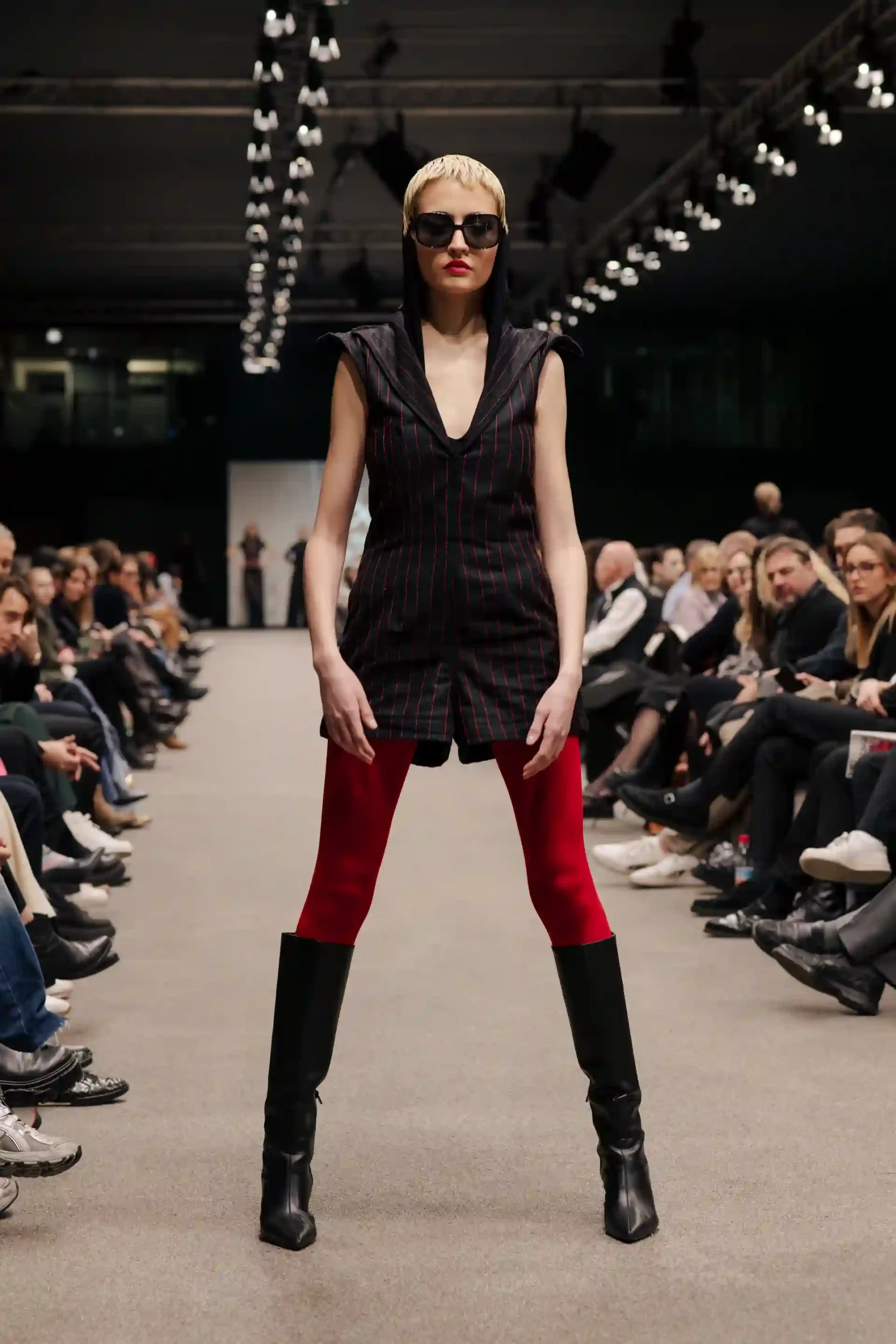 Elegantes Model mit kurzer Frisur und Sonnenbrille, roter Leggings, langen Stiefeln, kurzem Kleid und weitem Ausschnitt – sehr modern und cool.