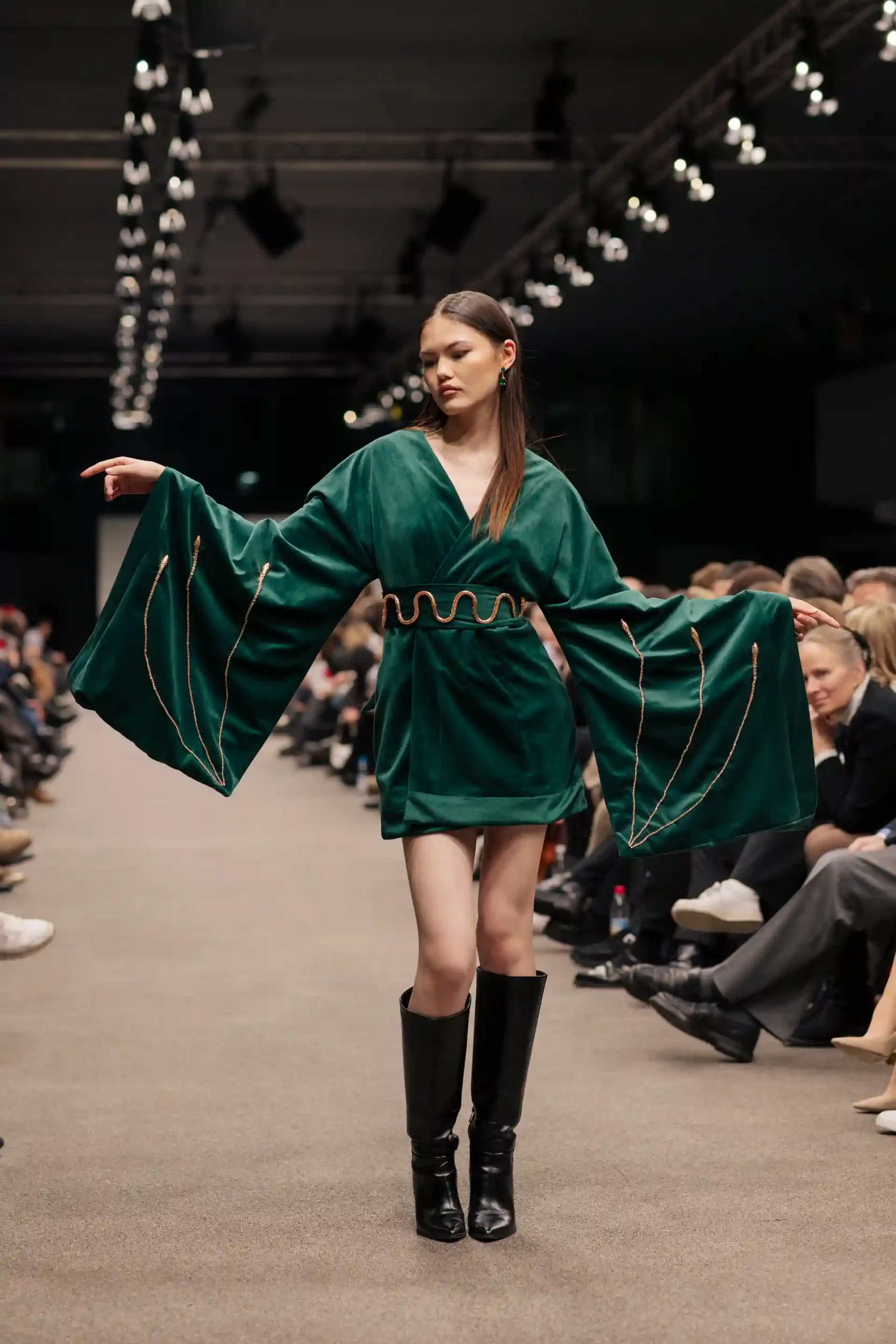 Das Modell steht auf dem Laufsteg, die Arme ausgestreckt, und trägt ein grünes Velourskleid mit markanten Ärmeln und einem dekorativen gold-braunen Gürtel uns schwarzen Stiefeln.