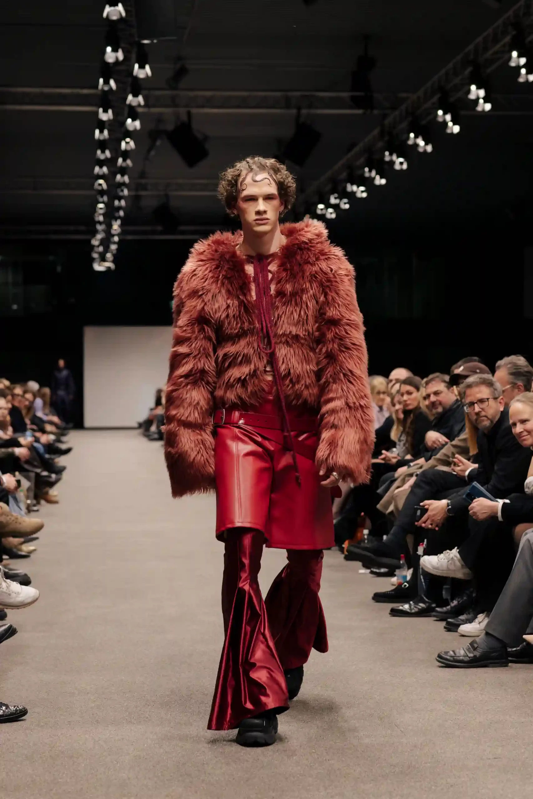 Ein männliches Model mit geschminktem Gesicht trägt ein auffälliges Outfit aus rotem Fell und Leder in kupferroter Optik. Ein sehr ausgefallenes, ungewöhnliches Erscheinungsbild.