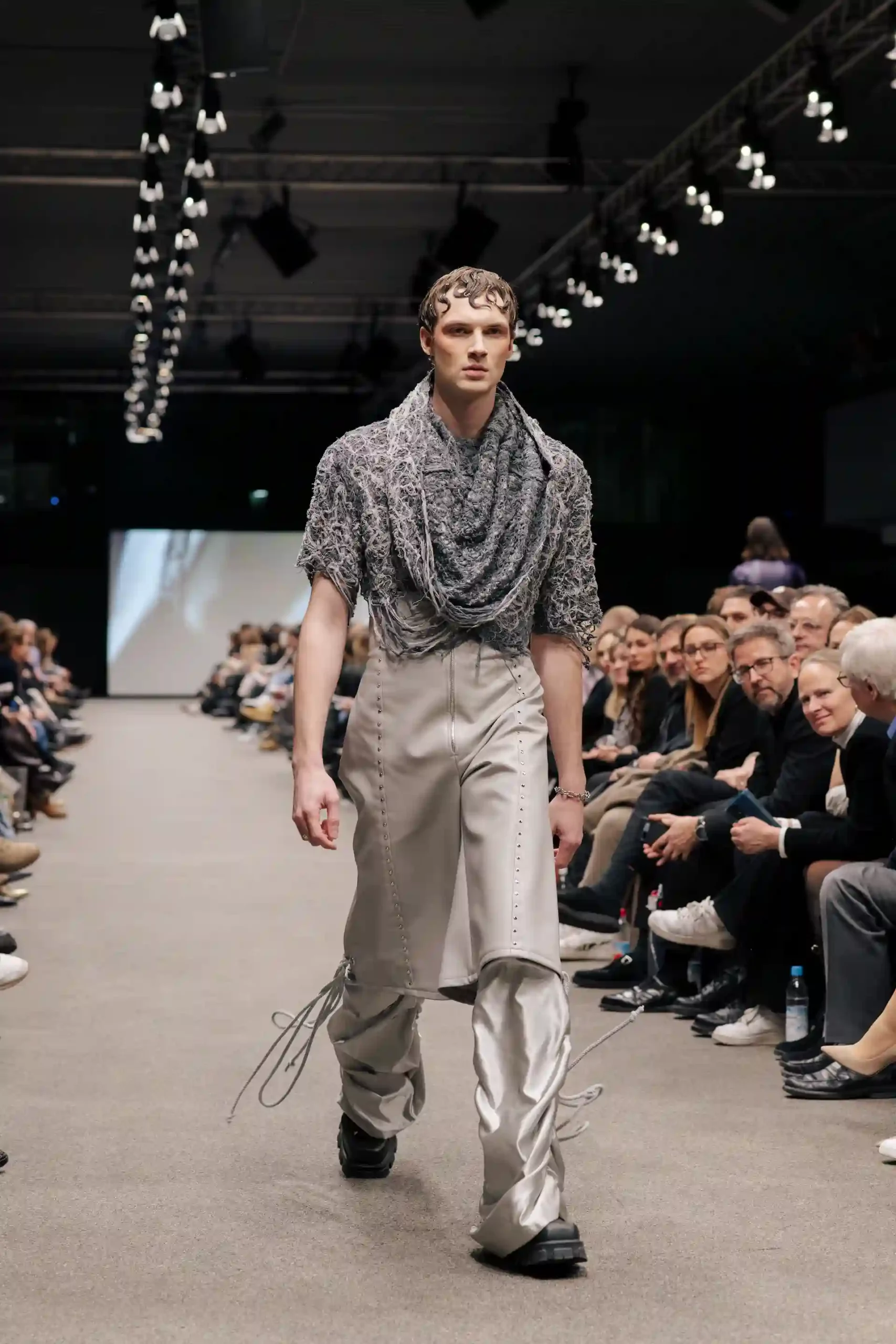 Ein männliches Modell zeigt ein Ensemble aus Leder und Stoff: ein graues Netzoberteil, eine stylische Lederhose und eine auffällige, ungewöhnliche Hose.