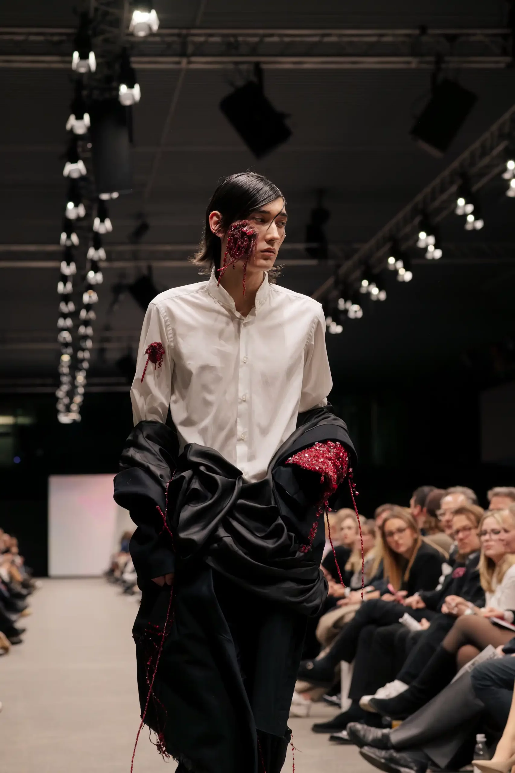 Herrenmodell mit weißem Hemd und schwarzer Hose, mit roten Steinakzenten in der Kleidung und einem Bereich auf der linken Gesichtshälfte, der kunstvoll mit kleinen roten Steinen drapiert ist.