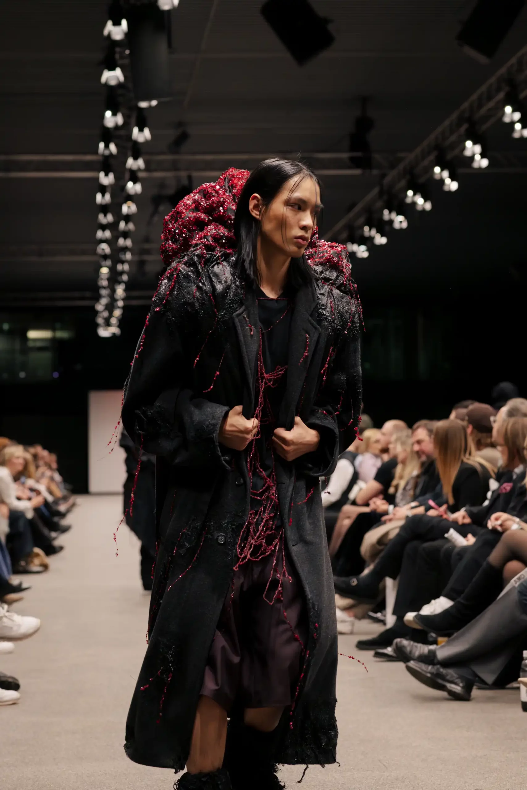 Herrenmodell in schwarzer Kleidung mit langem Mantel, in den zahlreiche feine Steinstränge eingearbeitet sind. Auf dem unteren Teil des Mantels befindet sich eine große rote Fläche aus vielen kleinen Steinen, drapiert wie ein Rucksack oder eine Kapuze.