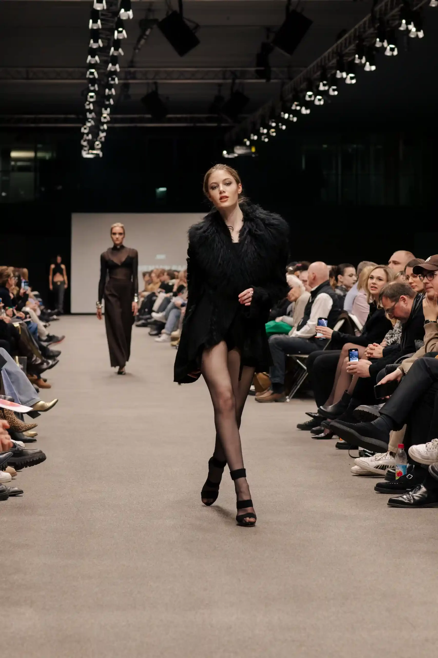 Model auf dem Laufsteg, trägt einen eleganten schwarzen Mantel mit seitlichen Federn, lange Beine in schwarzen Nylons und schwarze Stöckelschuhe, schreitet anmutig.