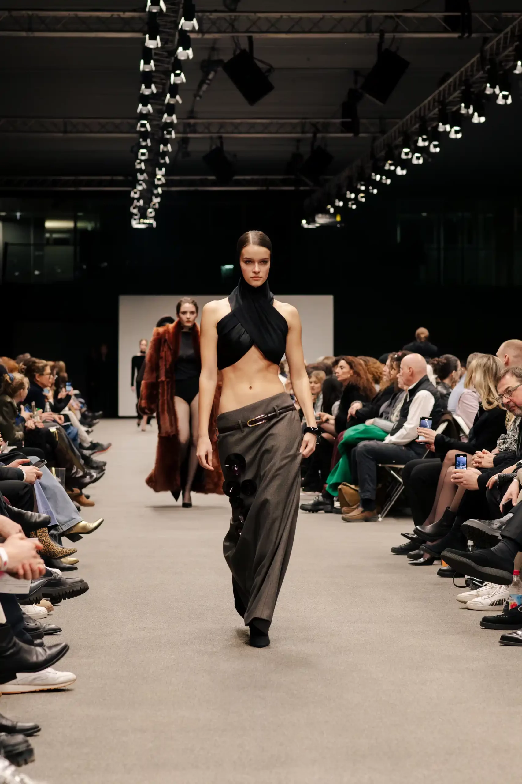 Model in stilvollem Outfit mit freiem Bauchnabel; hinter ihr gehen weitere Models den Catwalk entlang, während die Zuschauer seitlich gespannt beobachten.