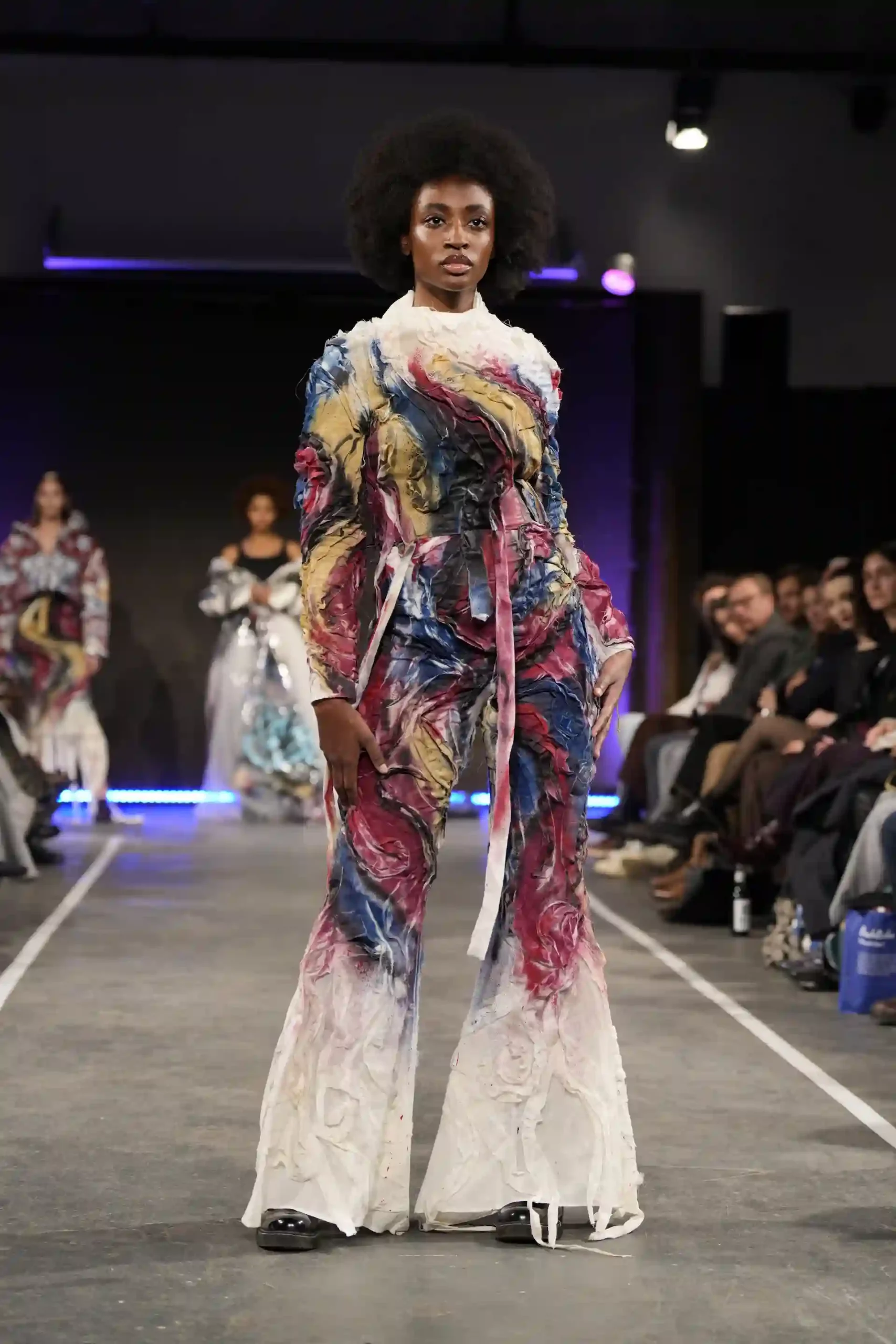 Ein People-of-Color-Model trägt ein buntes Outfit auf dem Laufsteg, das Publikum ist begeistert.