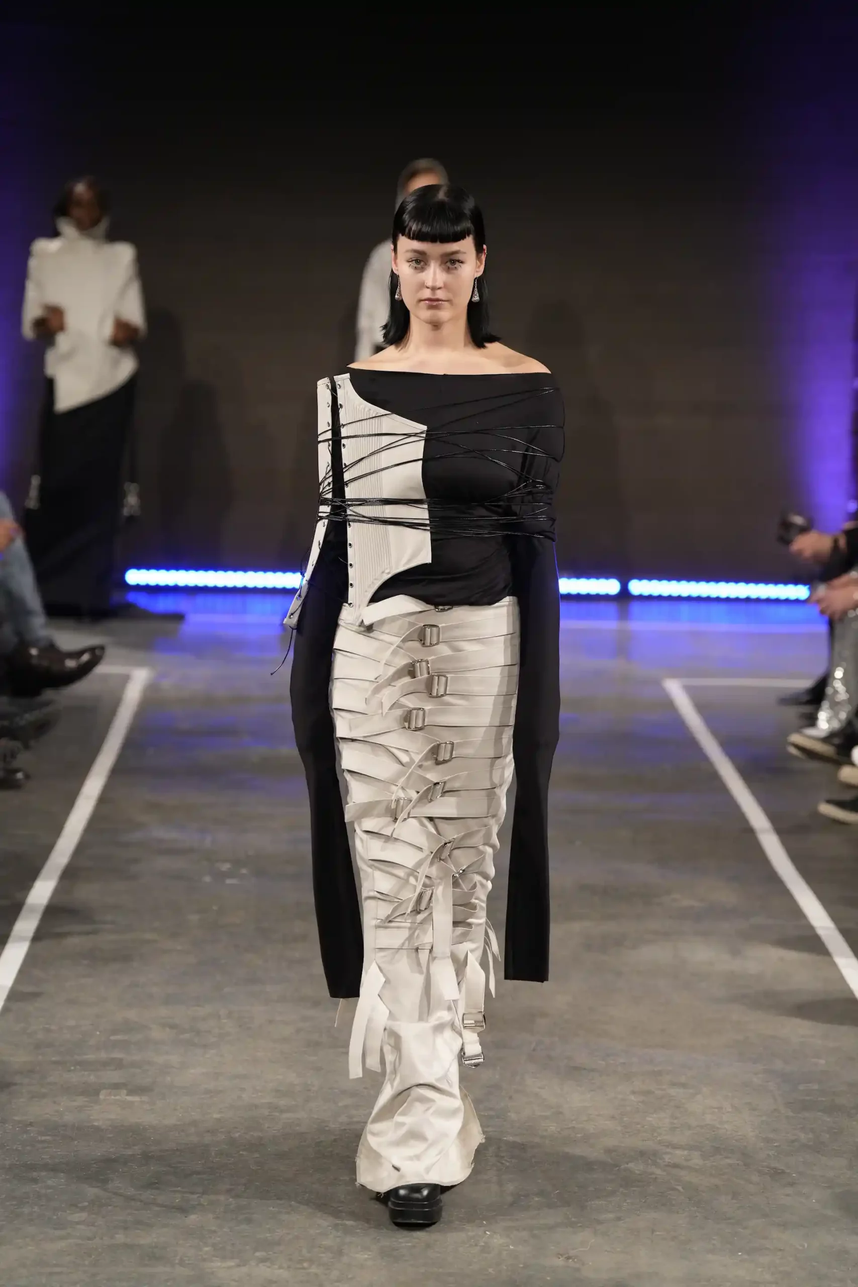 Ein Model auf dem Laufsteg trägt eine einzigartige schwarz-weiße Silhouette mit raffinierten Bindungen und einer Art Riemen um die Brust. Wie aus einem Science-Fiction-Film.