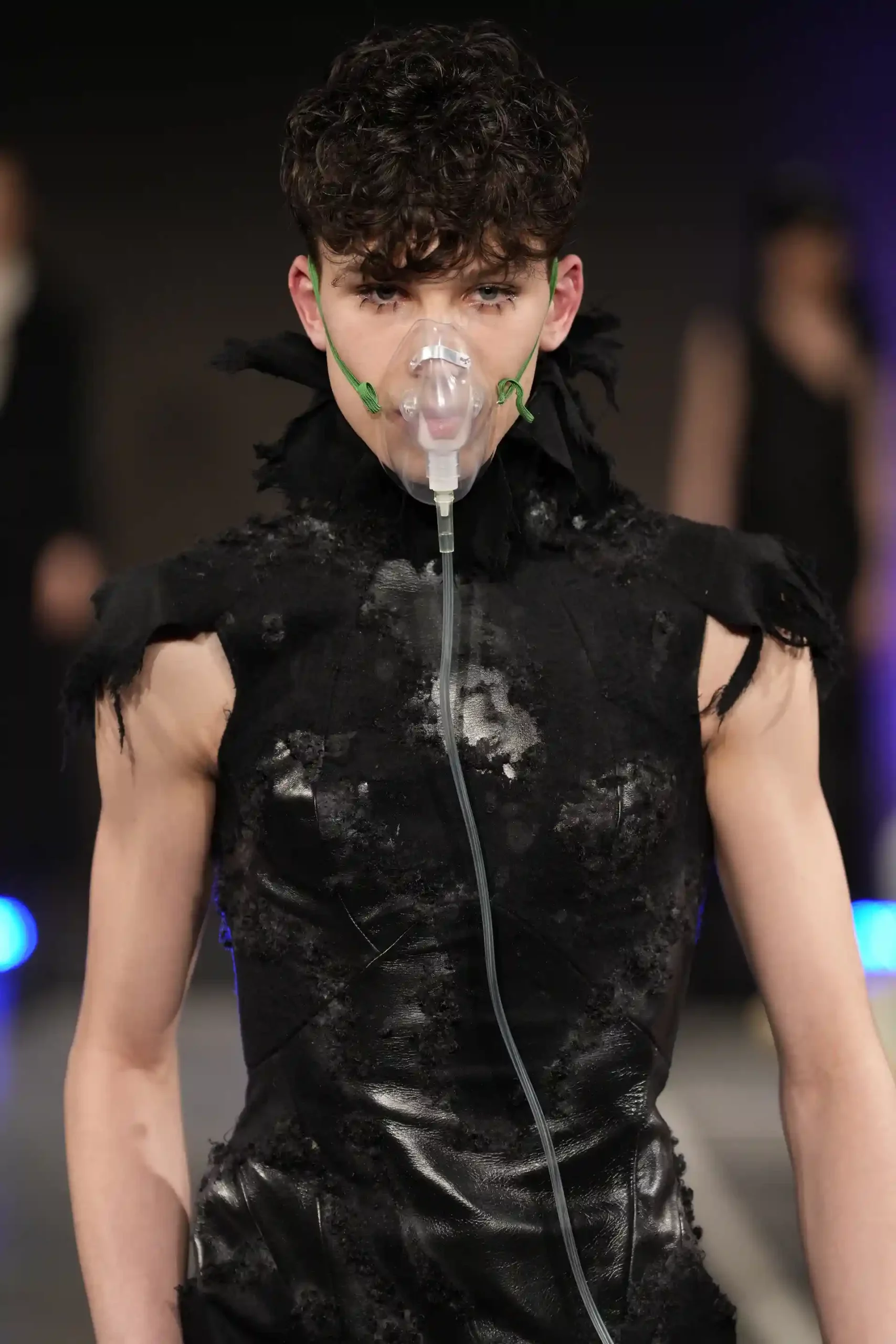 Ein Model in schwarzer, glänzender Lack- und Lederkleidung mit Atemschutz, überbetonten Augenbrauen, Oberteil mit freien Armen.