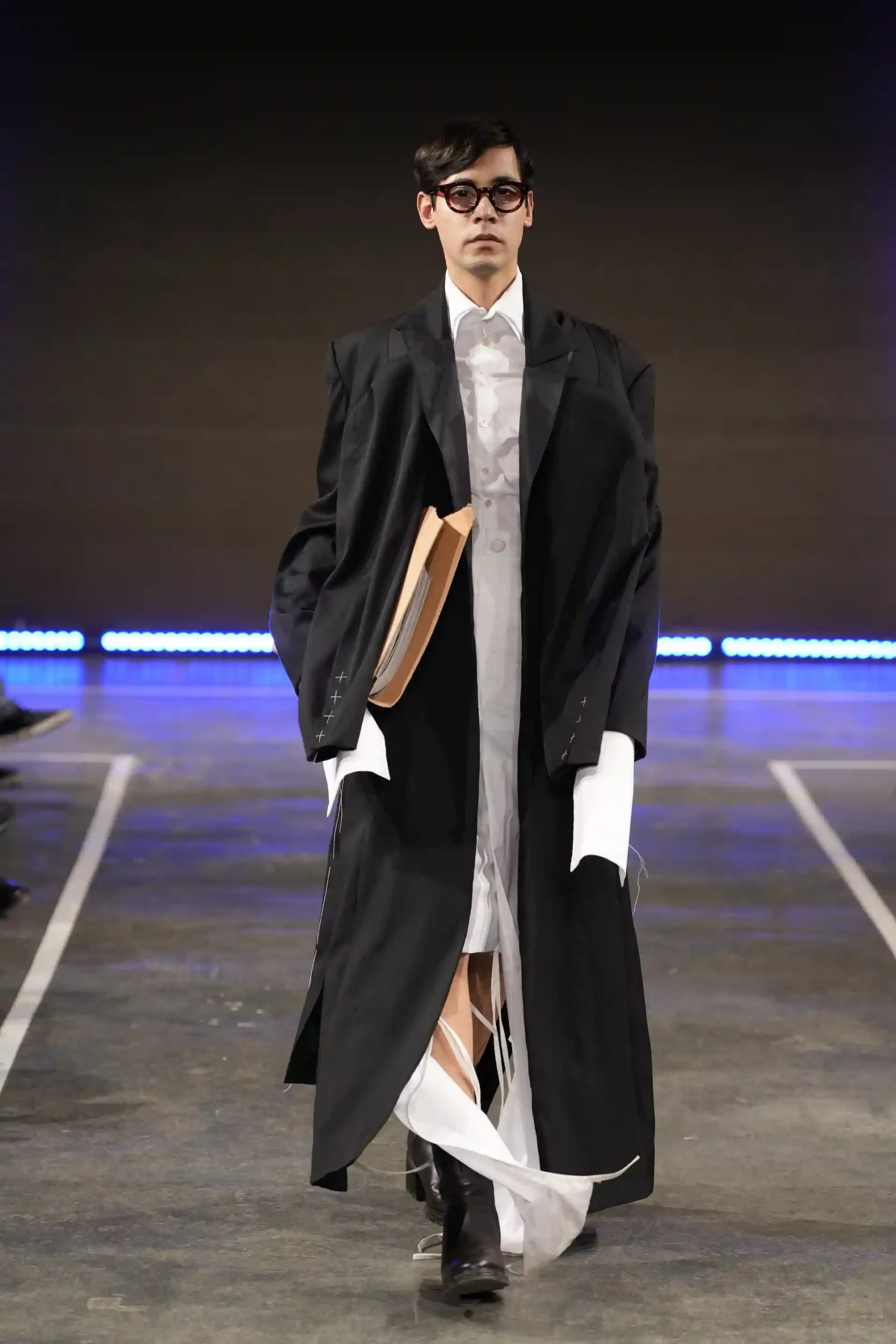 Ein avantgardistisches Model mit runder schwarzer Brille trägt einen schwarzen Mantel und ein weißes Hemd – jedoch keine Hose. Wie ein Anwalt aus der Zukunft auf dem Laufsteg.