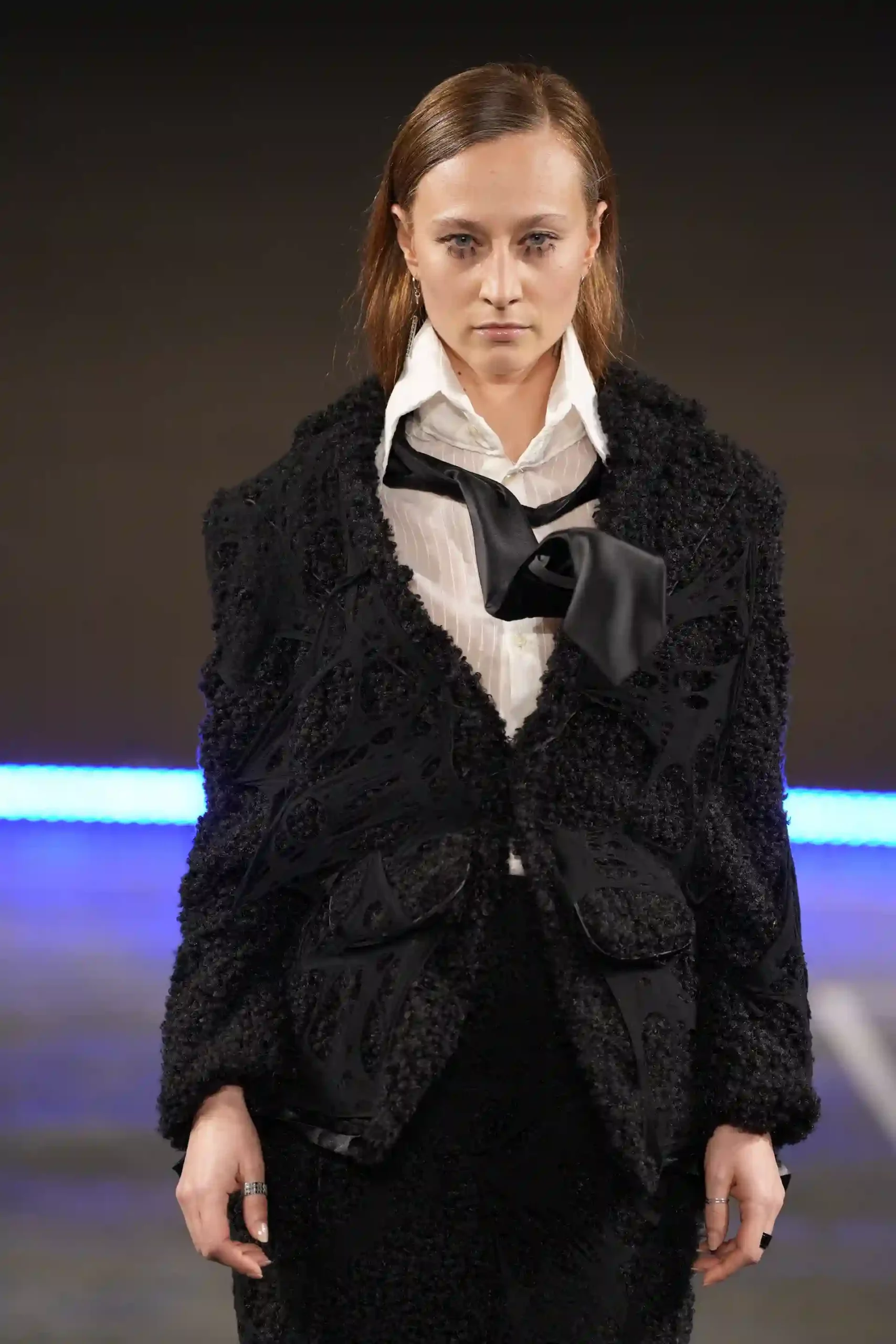 Ein Modell trägt eine schwarze Wolljacke, ein weißes Hemd und eine glänzende schwarze Krawatte, die auffällig nach oben gesteckt ist.