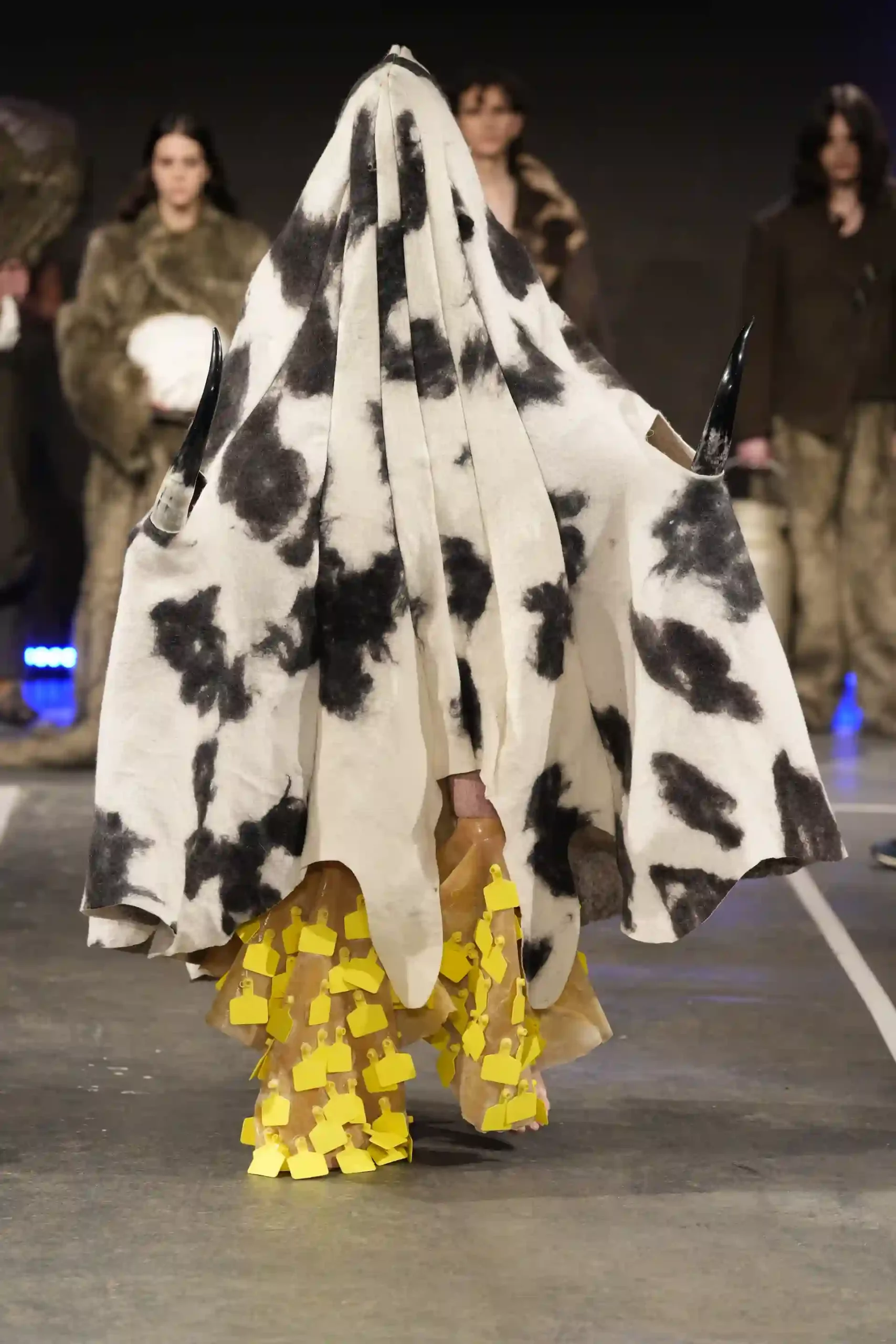 Model mit außergewöhnlichns Outfit, mit Hörnern und einem gemusterten Umhang auf dem Laufsteg.