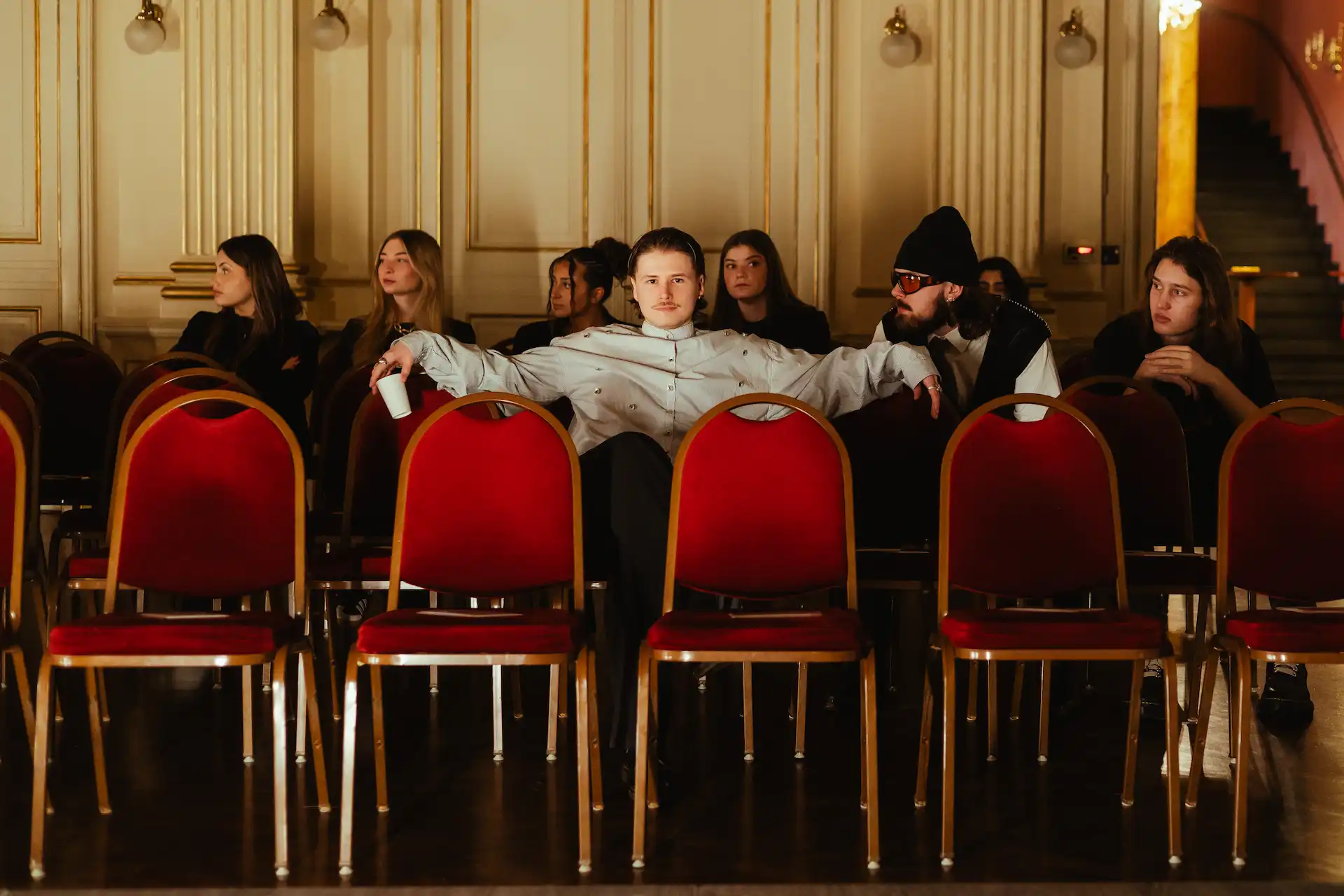 Eine Gruppe von Modedesign-Studierenden der AMD sitzt auf roten Stühlen im Palais und blickt gespannt zur Bühne, während sie auf die Modenschau warten.