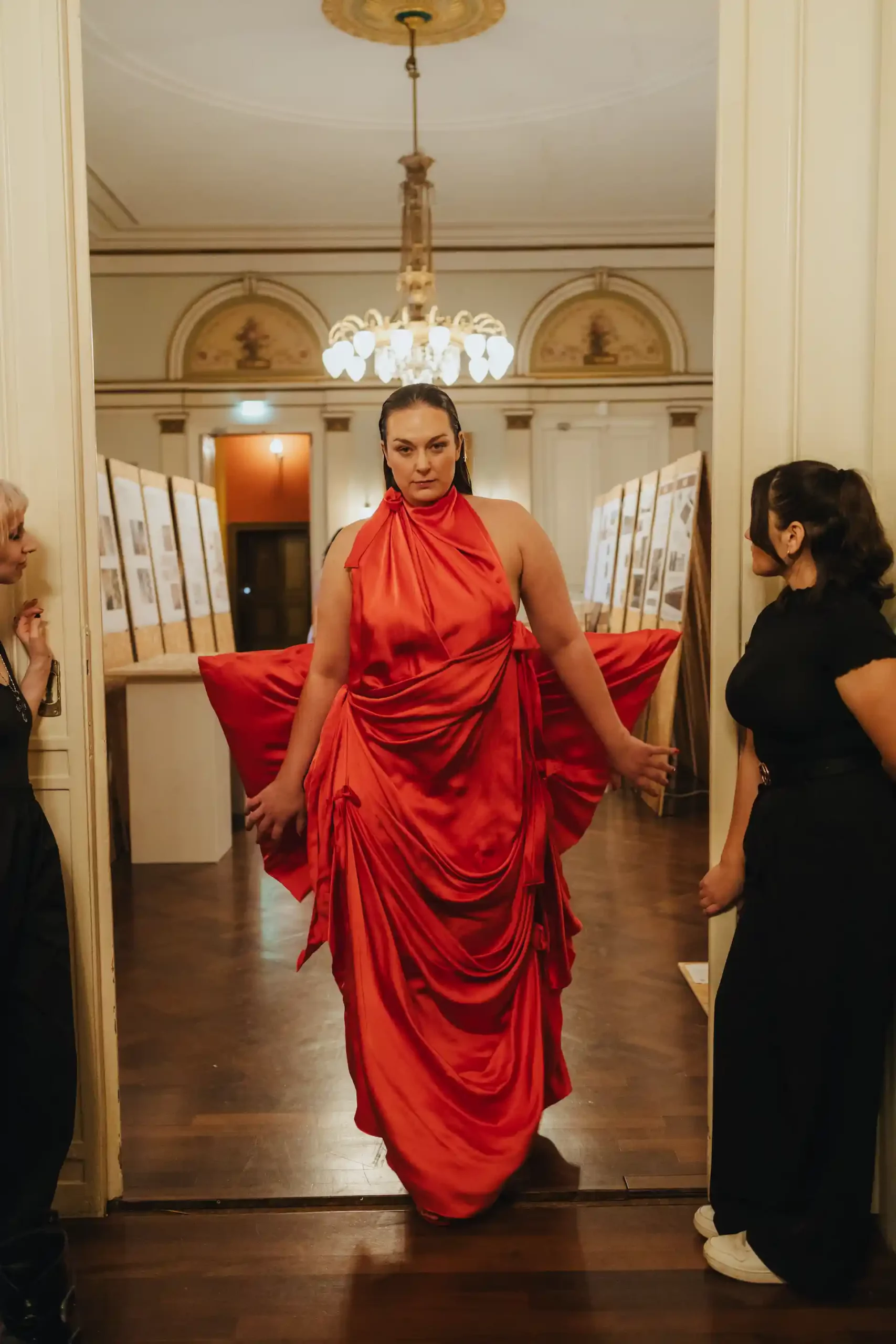 Ein Model backstage bei der Anprobe eines auffälligen roten Abendkleides; rechts und links stehen die Designer und helfen dabei.