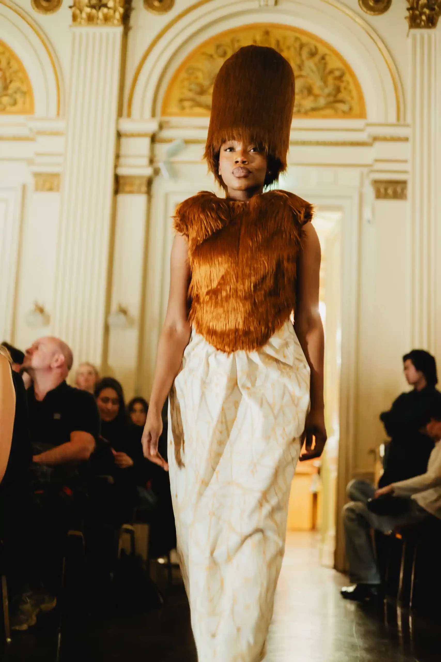 Ein People-of-Color-Model mit einem extravaganten, überdimensional großen Hut aus braunem Fell, ebenso wie das Oberteil.