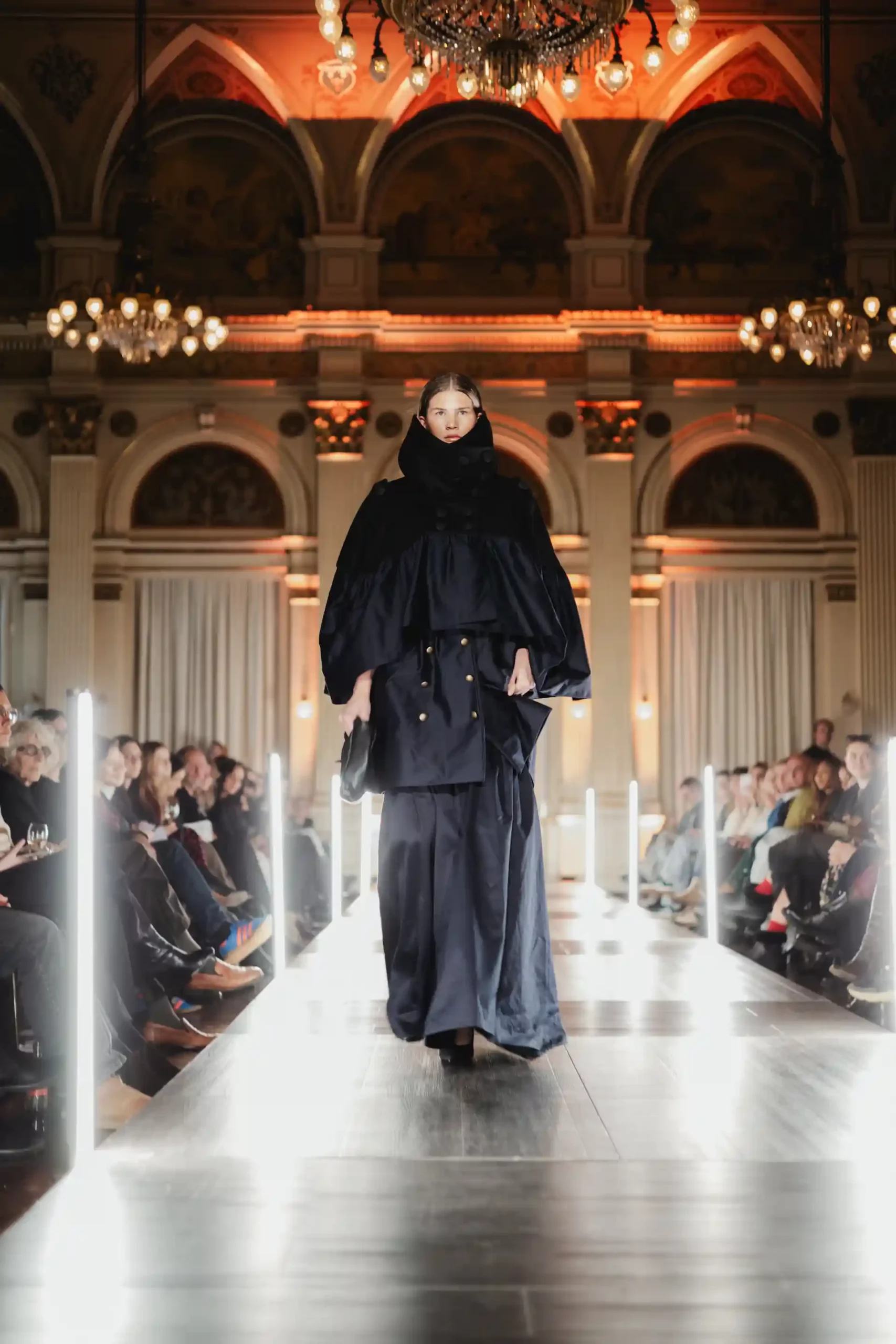 Ein Model in moderner, schwarzer Garderobe schreitet über den glanzvollen Laufsteg mit hohen Decken, funkelnden Kronleuchtern und einem begeisterten Publikum.