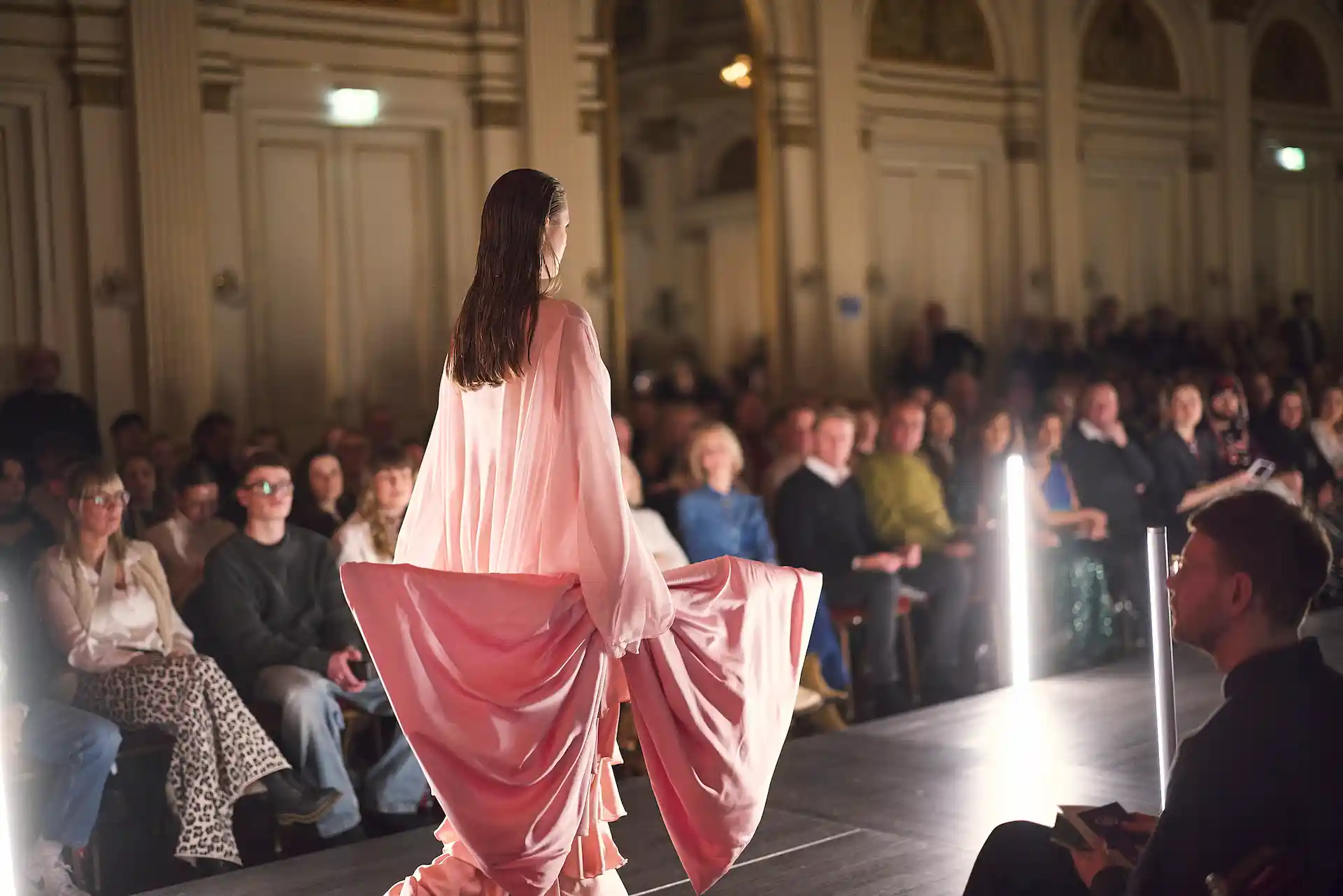 Model auf dem Catwalk in einem pinken Kleid mit Publikum im Hintergrund.