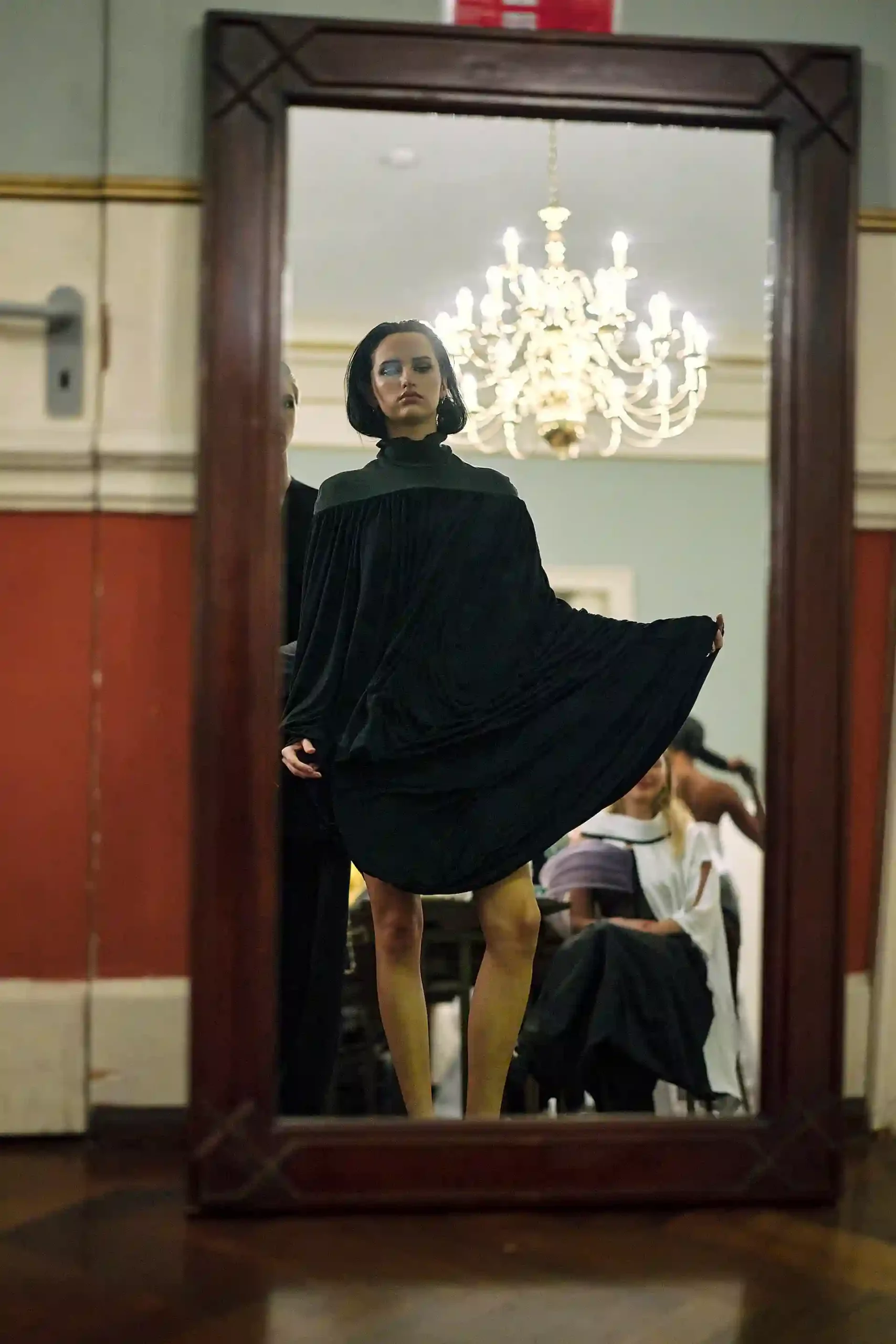 Backstage Moment: Model mit kurzem Haarschnitt trägt ein schwarzes Kleid und betrachtet sich in einem großen Spiegel.