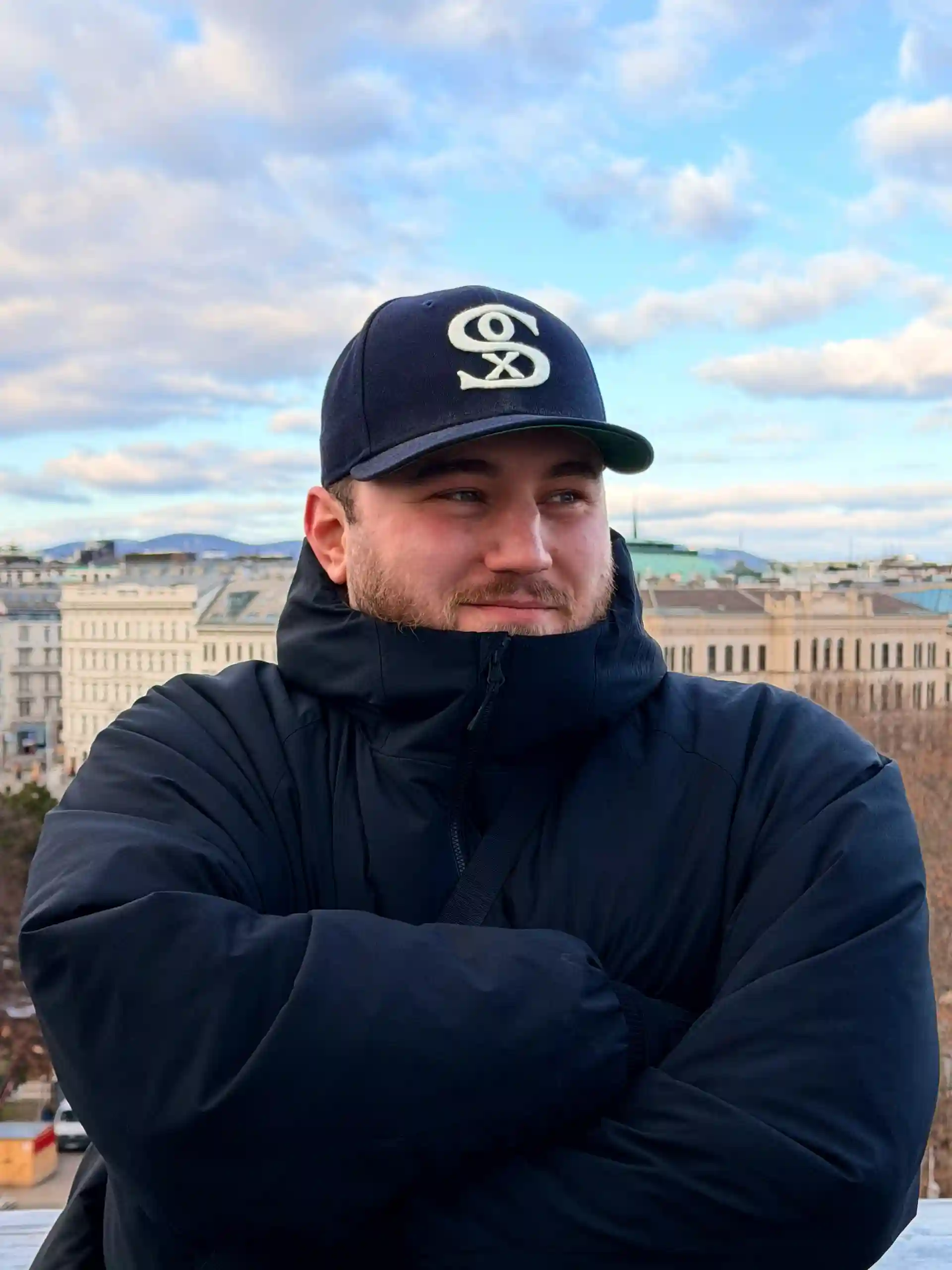 Porträt von Samet Osman mit Cap und dunkler Jacke, der auf einer Terrasse mit verschränkten Armen posiert.