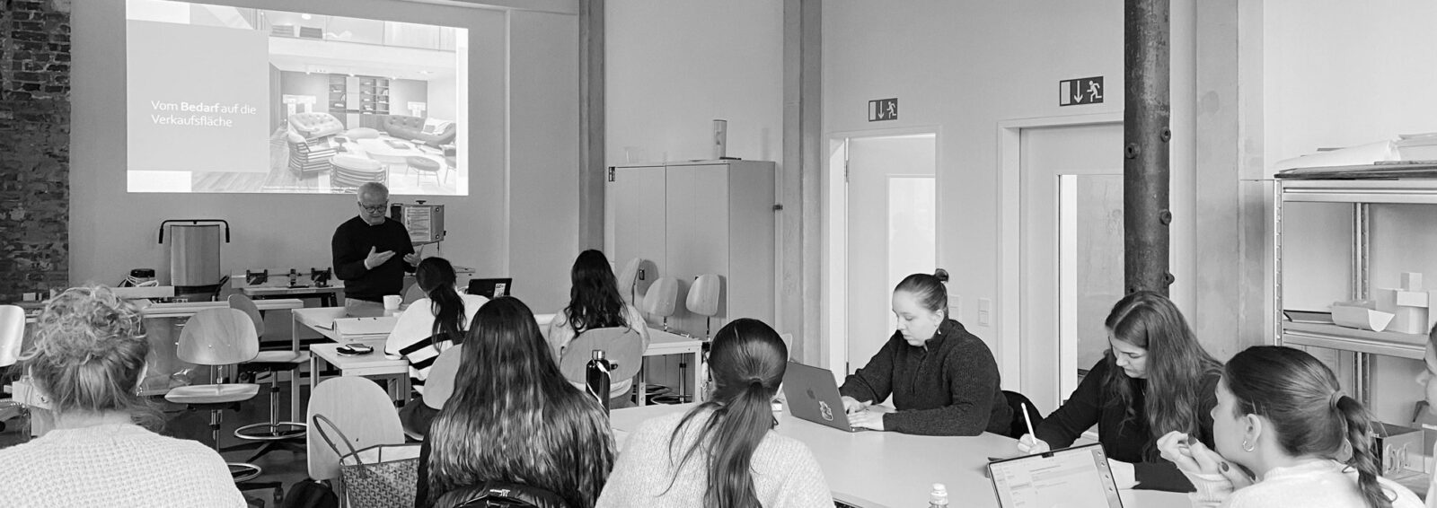 Martin Aichberger hält seinen Vortrag vor einer sitzenden Gruppe angehender Interior Designer:innen in einer modernen Lernumgebung. Die Teilnehmer:innen arbeiten an Laptops, während im Hintergrund eine Diaschau präsentiert wird.