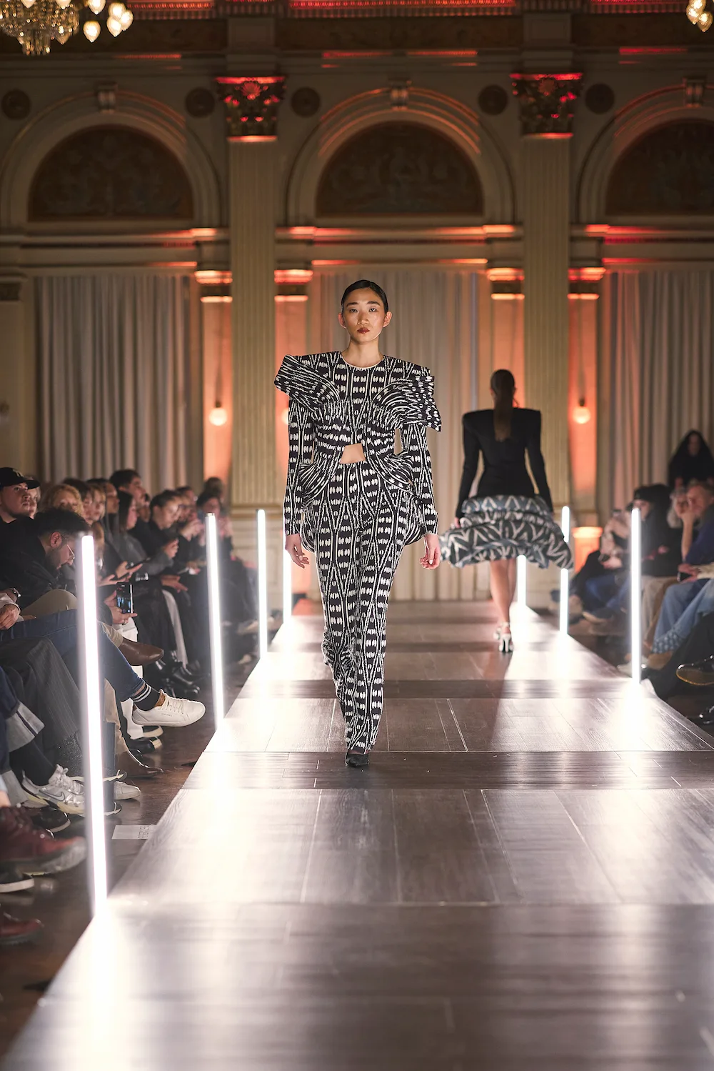 Catwalk mit beleuchteten Elementen neben der Bühne, während ein Model in stilvoller Kleidung läuft.