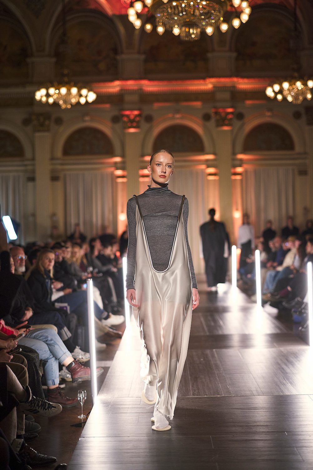 Publikum schaut einer Modenschau zu, während ein Model mit futuristischer Kleidung den Laufsteg betritt